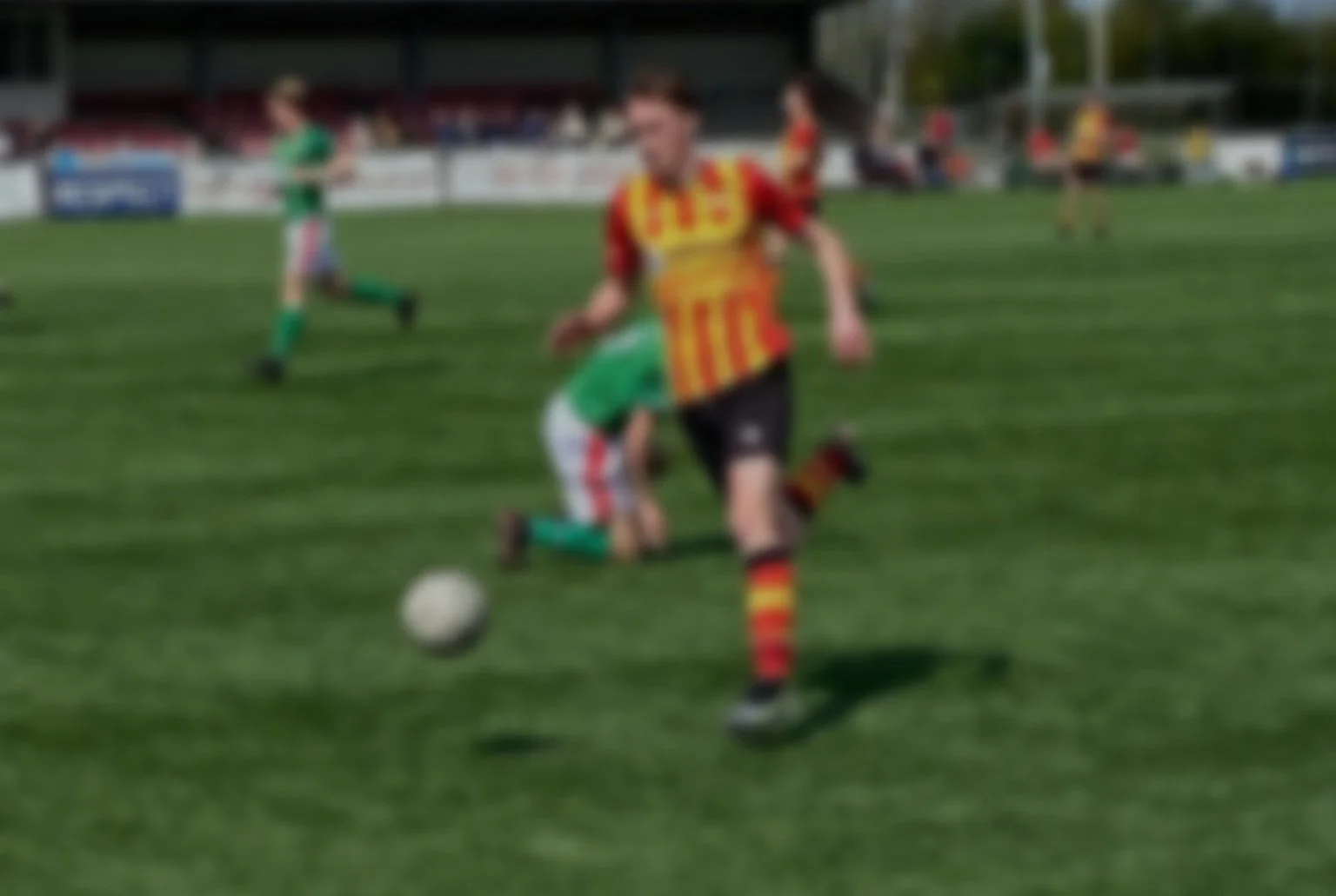 Een jeugdvoetbalwedstrijd met spelers in groene en rode shirts op een grasveld, een speler in een rood-geel gestreept shirt wordt gevolgd door een speler in groen.