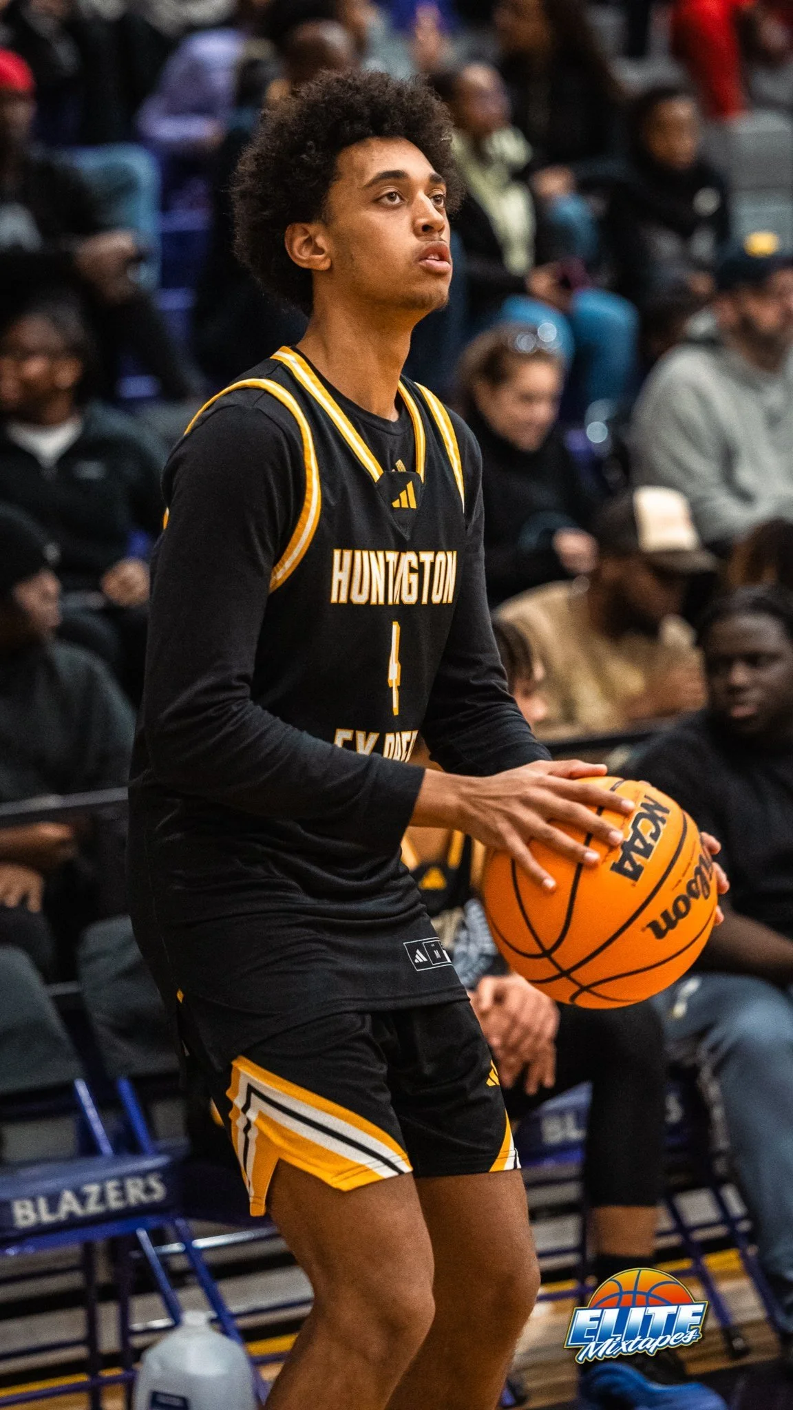 Een man in een zwart en geel basketbaluniform met 'Huntington' erop, die met beide handen een basketbal vasthoudt tijdens een basketbalwedstrijd.