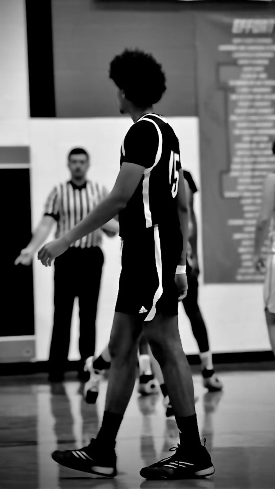 Zwart-wit foto van een indoor volleybalwedstrijd met een speler in zwarte sportkleding en andere spelers en een scheidsrechter op de achtergrond.
