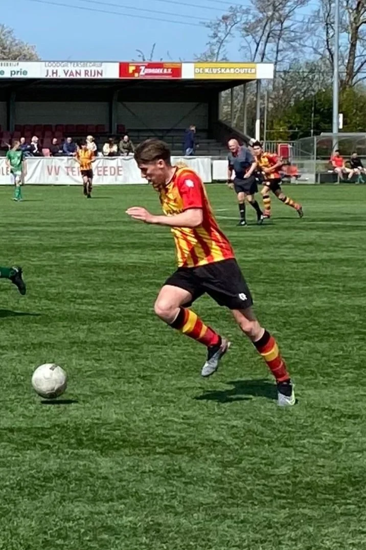 Een jongen speelt voetbal op een grasveld, gekleed in een rood en geel gestreepte voetbaloutfit, met mensen op de achtergrond en een stadiontribune.