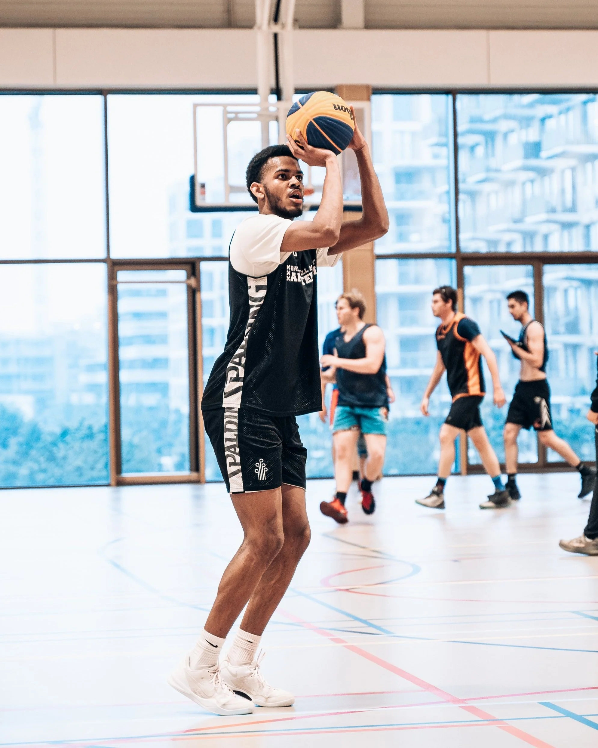 Een man die basketbal speelt in een sporthal met grote ramen, terwijl andere mensen trainen op de achtergrond.