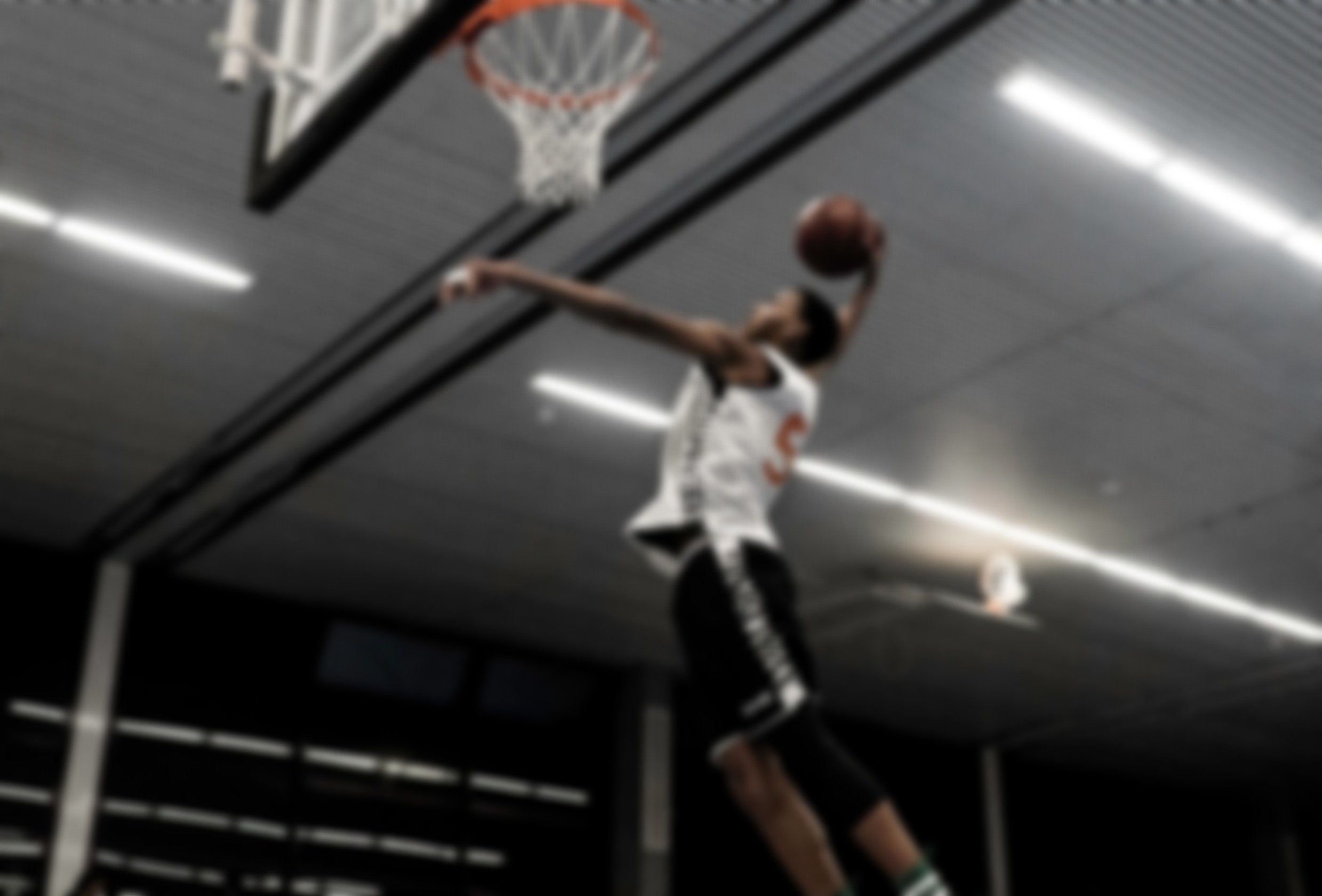 Een jongen speelt basketbal in een binnensporthal, springt omhoog met de bal richting de basket, op de achtergrond is het plafond en verlichting zichtbaar.