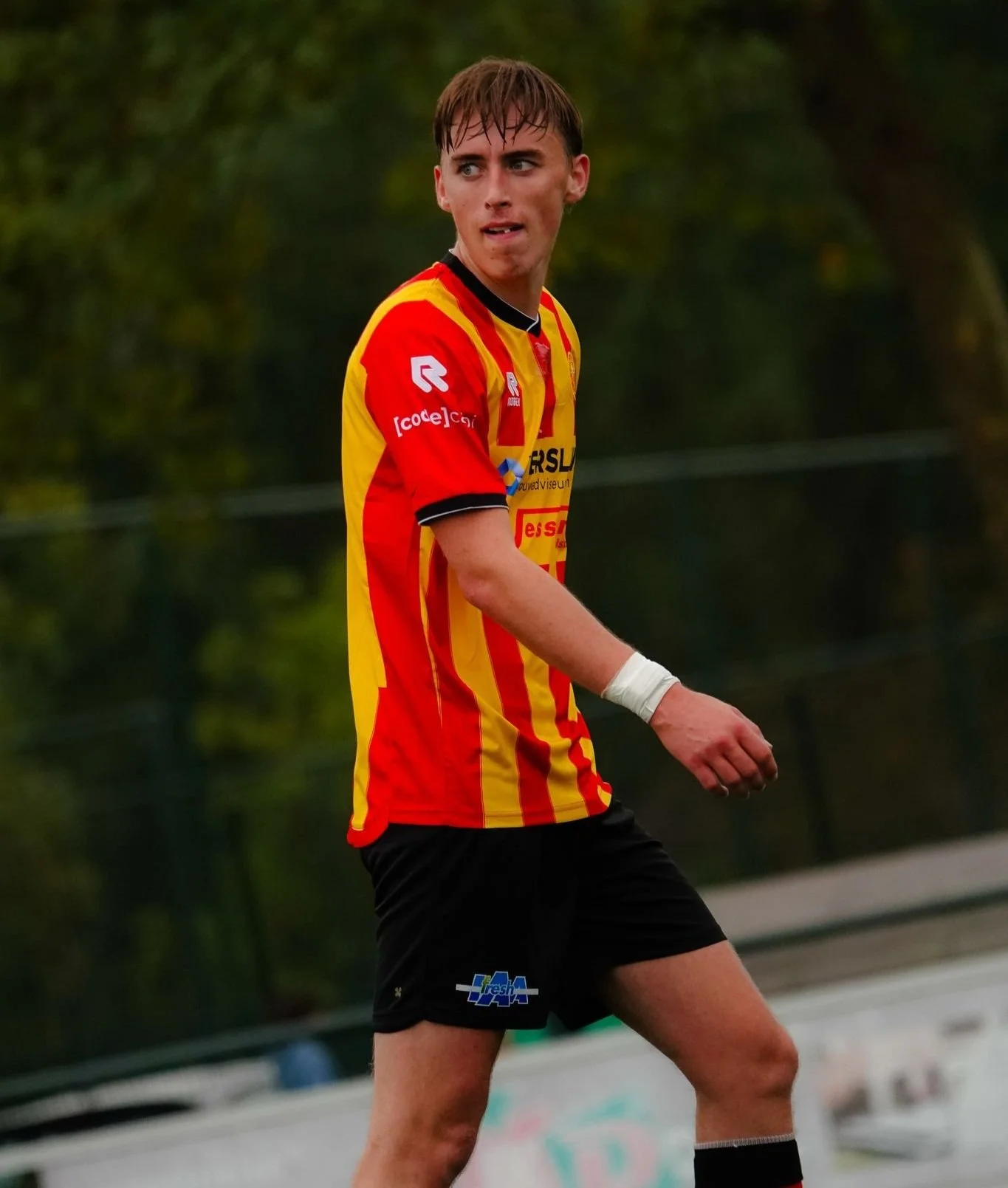 Een jonge man in een rood en geel sportshirt en zwarte joggingbroek op een sportveld, mogelijk tijdens een wedstrijd of training, met bomen op de achtergrond.