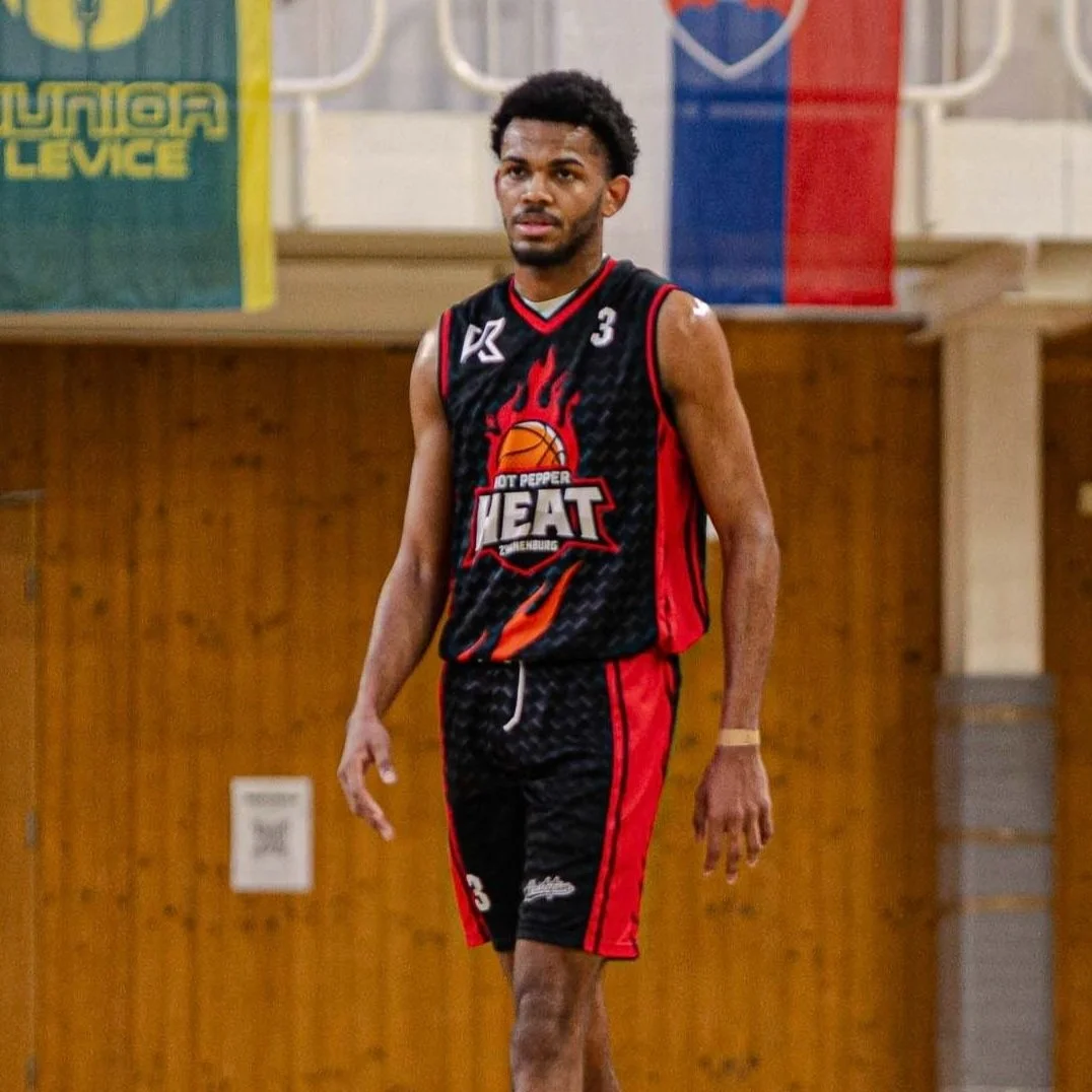 Een mannenbasketbalspeler in een zwart en rood teamuniform speelt op een basketbalveld in een sporthal.
