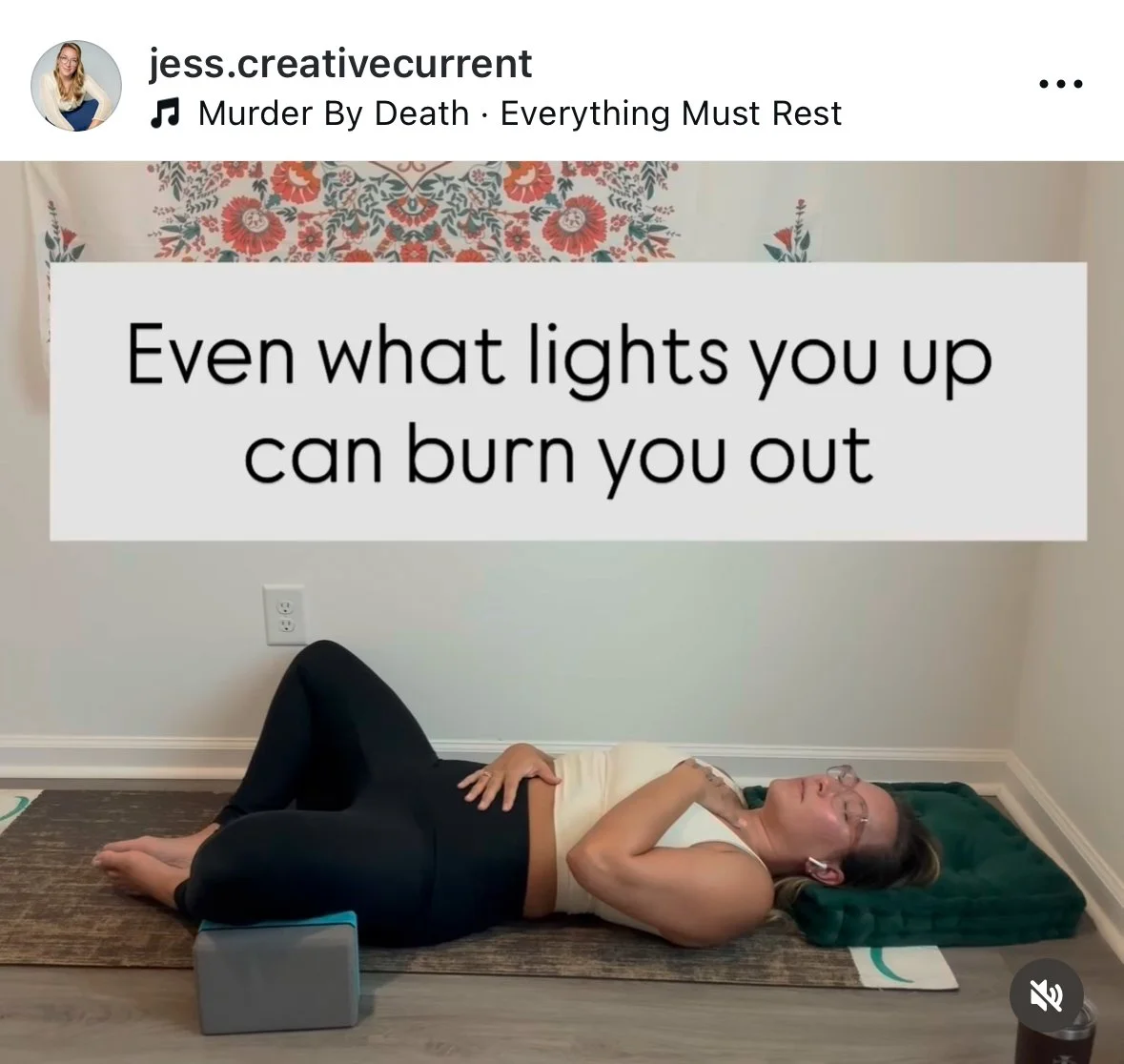 A woman lying on her side on a yoga mat, her head resting on a green pillow, with a blanket under her head and a block supporting her knees, smiling with her eyes closed.