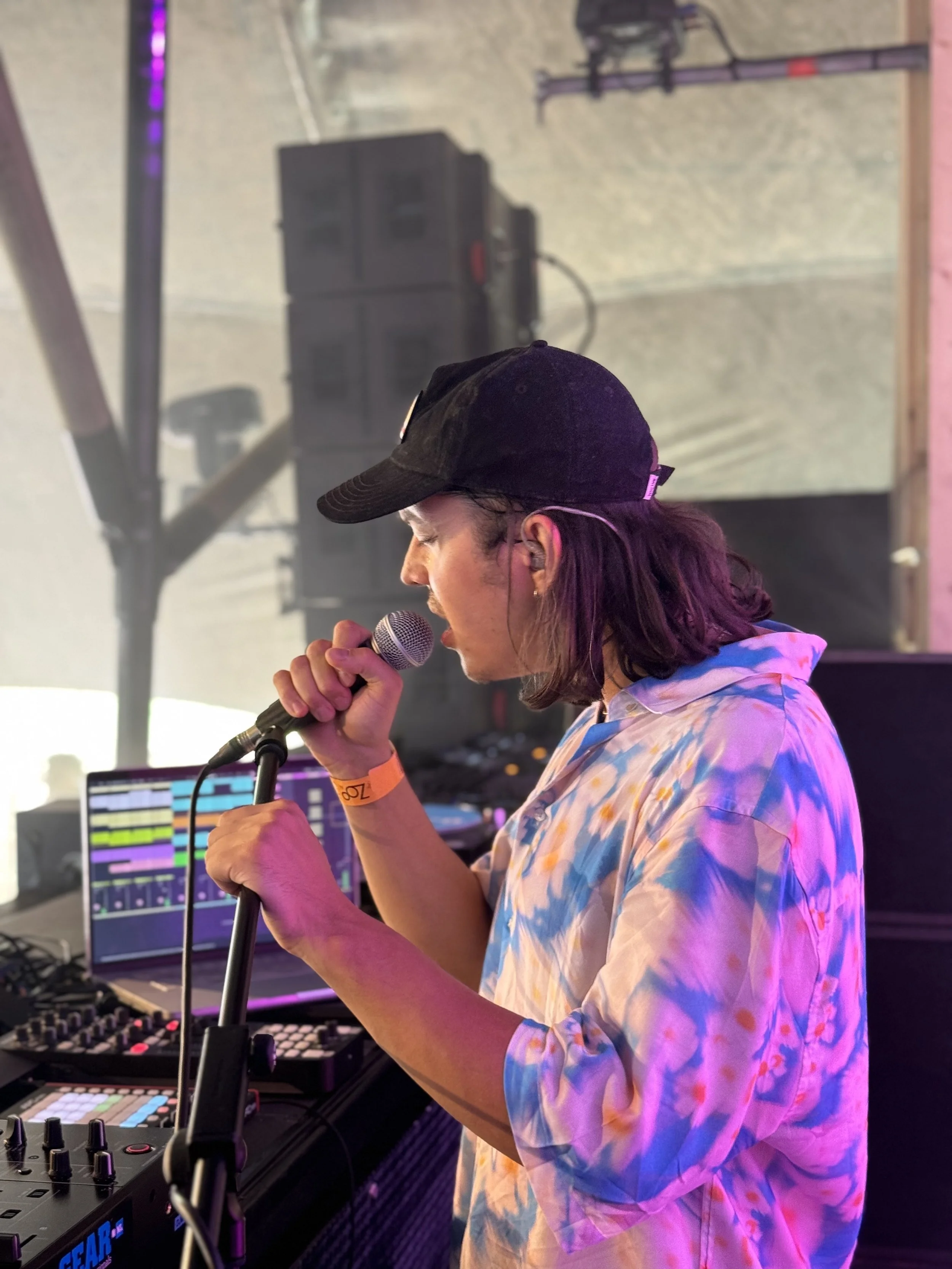 A person with long hair wearing a baseball cap and tie-dye hoodie, holding a microphone, appears to be DJing at an event with DJ equipment and a laptop in the background.