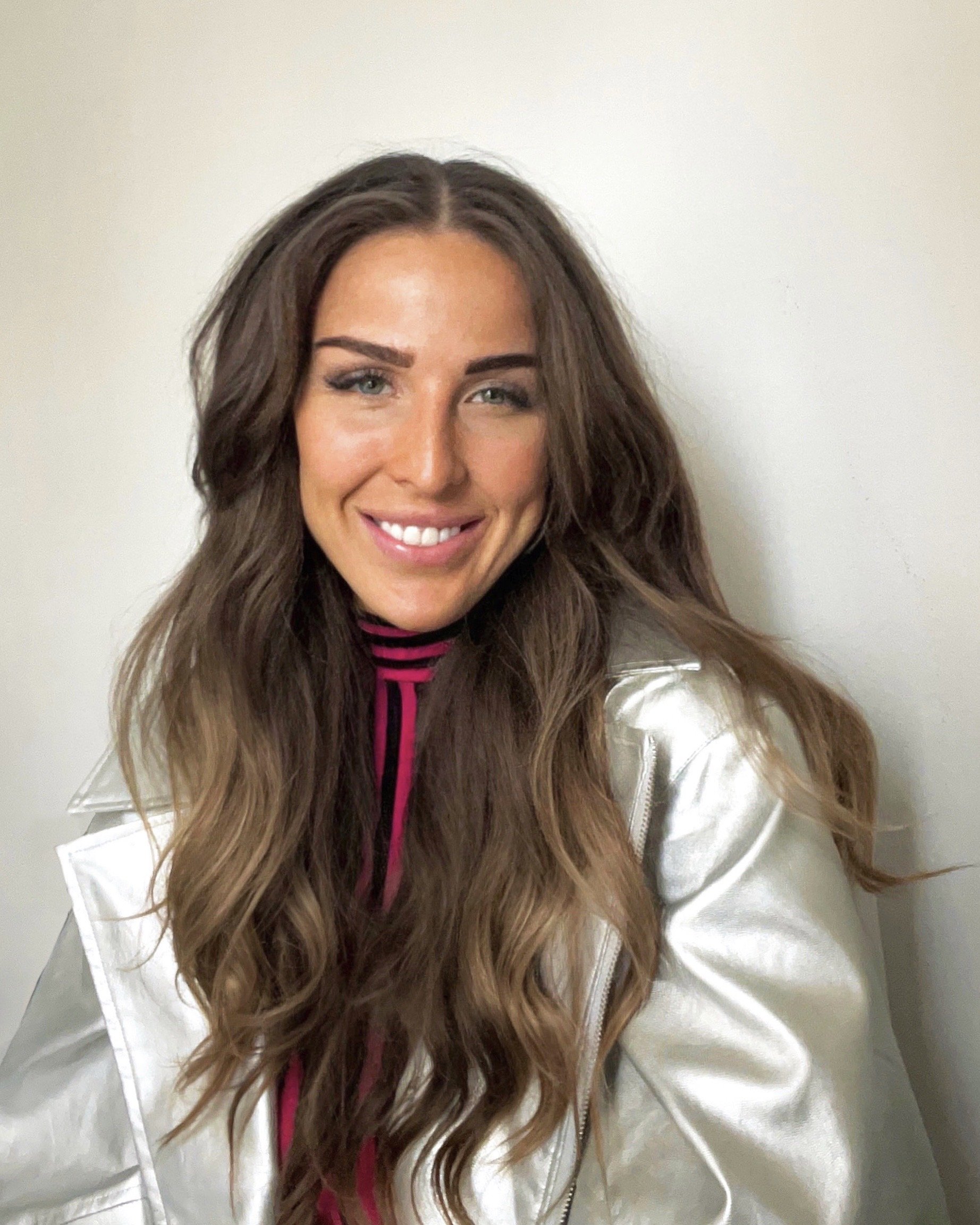 A woman with long wavy brown hair smiling, wearing a pink and black striped top and a light-colored jacket, standing against a plain wall.