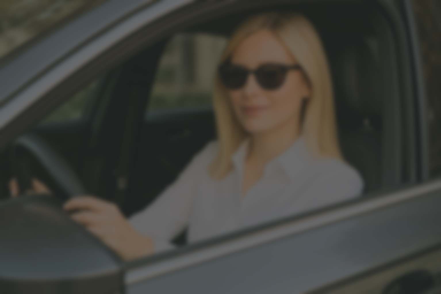 Een vrouw met blond haar en zonnebril zit achter het stuur van een auto, glimlachend.