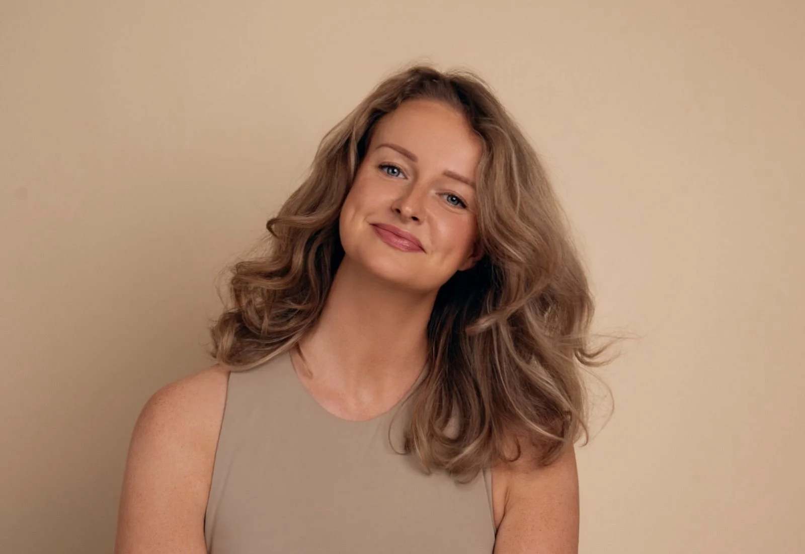 Portret van een lachende vrouw met golvend bruin haar tegen een beige achtergrond.