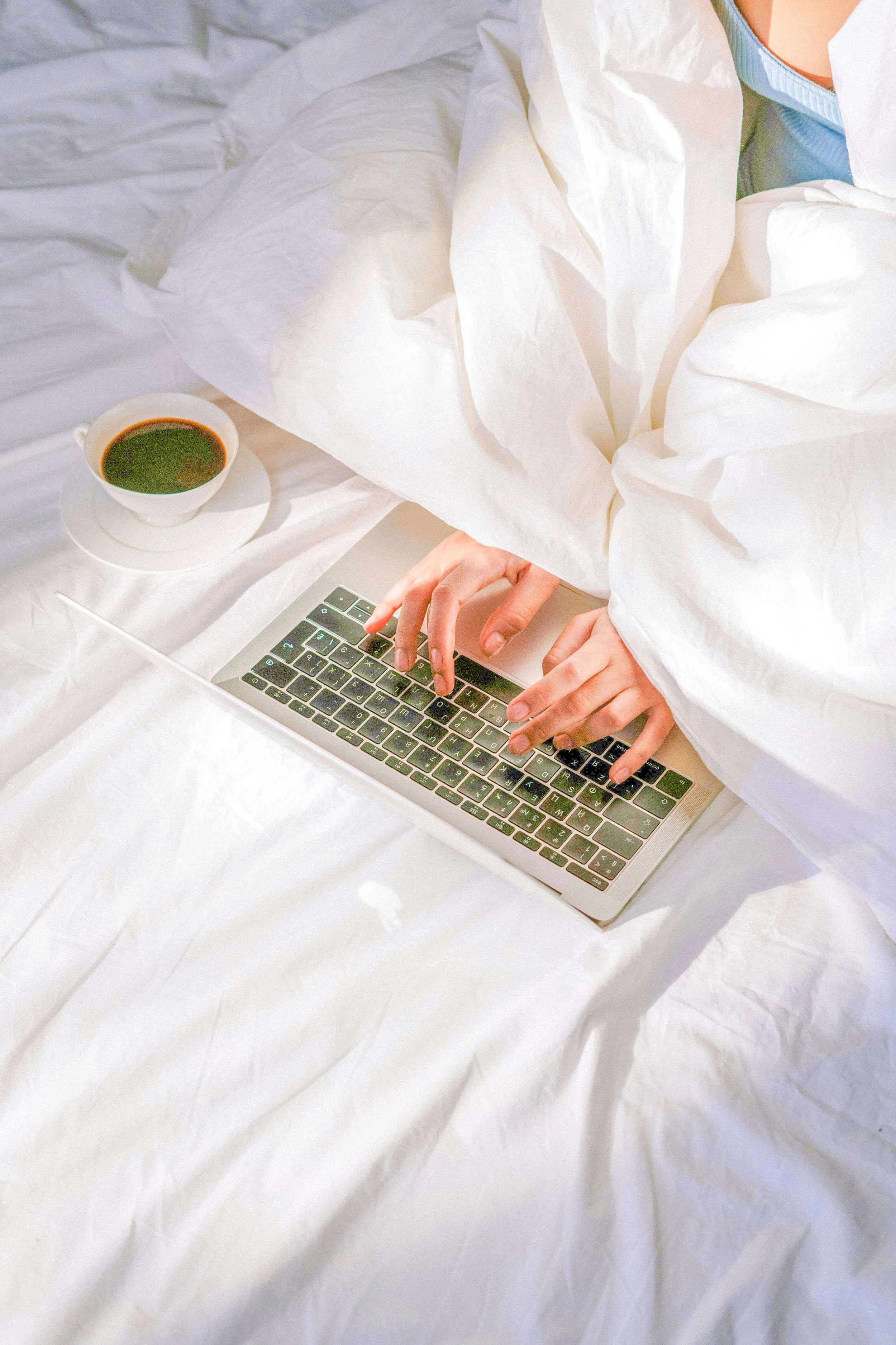 Une personne tape sur un clavier d'ordinateur portable blanc, avec une tasse de café ou de thé à côté, le tout posé sur une surface en tissu blanc.