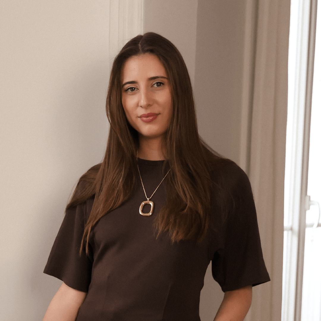 Jeune femme avec cheveux bruns, portant un vêtement marron et un collier, debout sur fond de mur blanc et une fenêtre avec rideaux.
