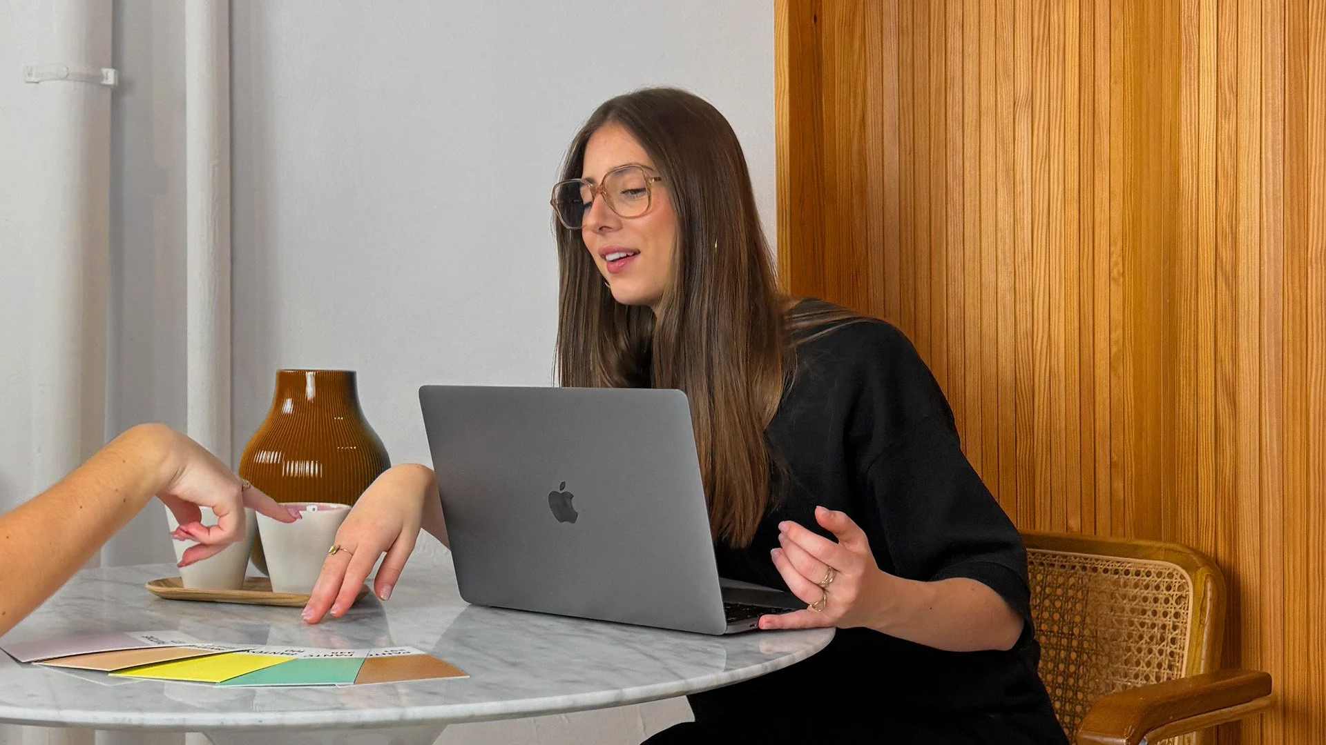Une femme avec des lunettes, assise à une table devant un ordinateur portable, en train de recevoir de petits objets d'une personne dont seule la main est visible.