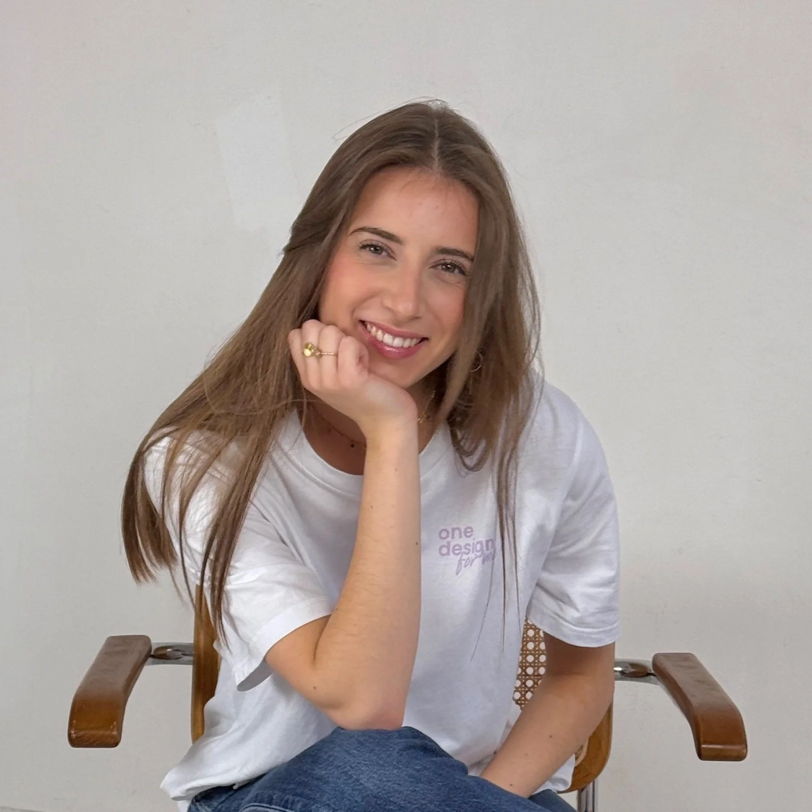 Une femme assise sur une chaise en bois, souriant, avec la tête reposant sur sa main, portant un t-shirt blanc et un jean, devant un mur blanc.