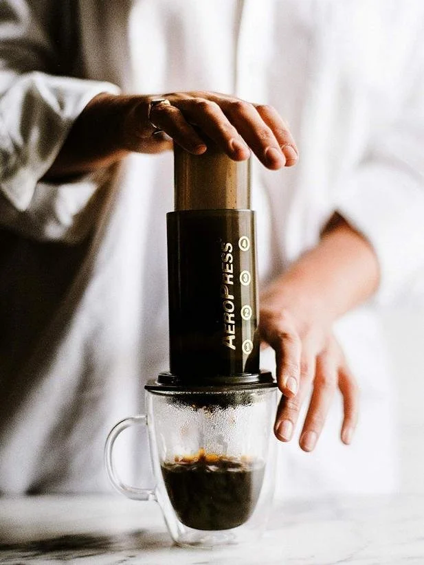 Person using AeroPress coffee maker to brew coffee into a glass mug.