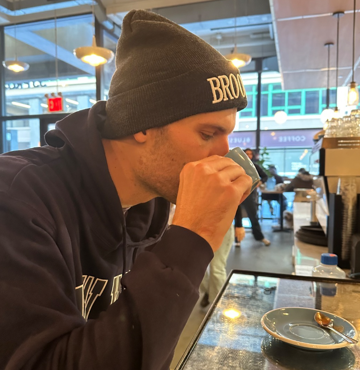 A man wearing a gray beanie and dark hoodie drinking from a light blue mug in a cafe, with a saucer and spoon on the table, and large windows in the background.