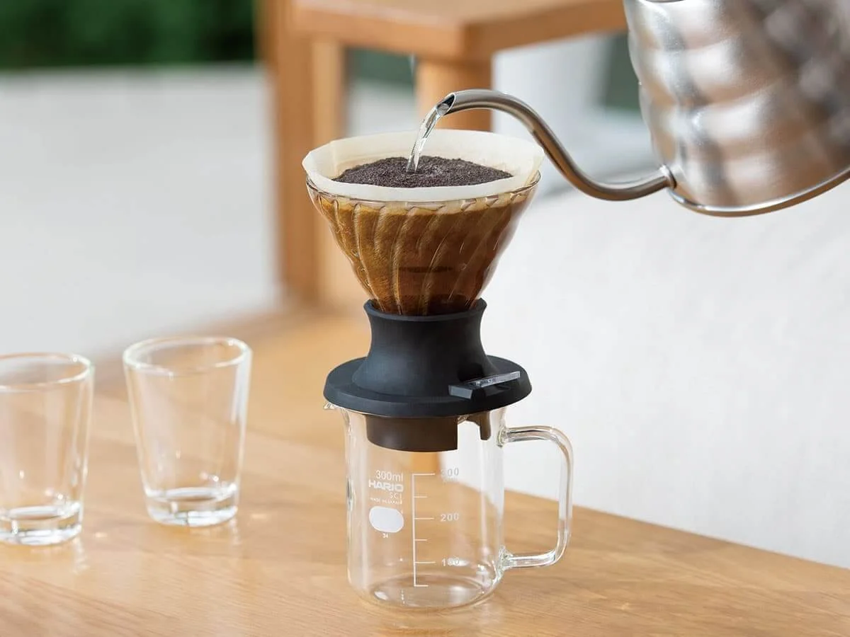 A pour-over coffee maker with ground coffee and hot water, set on a wooden table with two empty glasses nearby.