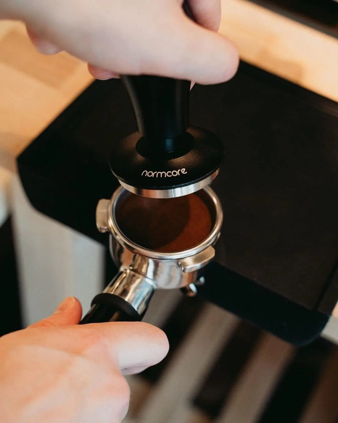 Close-up of a hand pressing a portafilter into a coffee tamper labeled 'normcore' on an espresso machine.