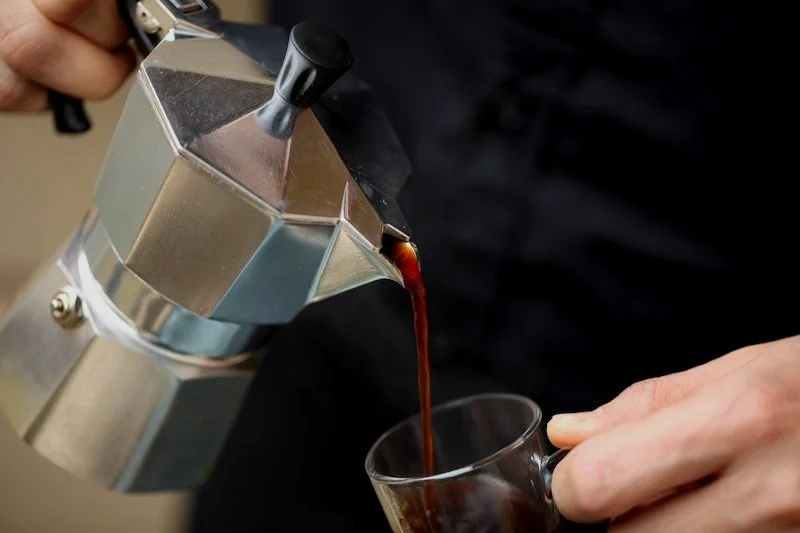 Person pouring hot brewed coffee from an espresso machine into a clear glass mug.