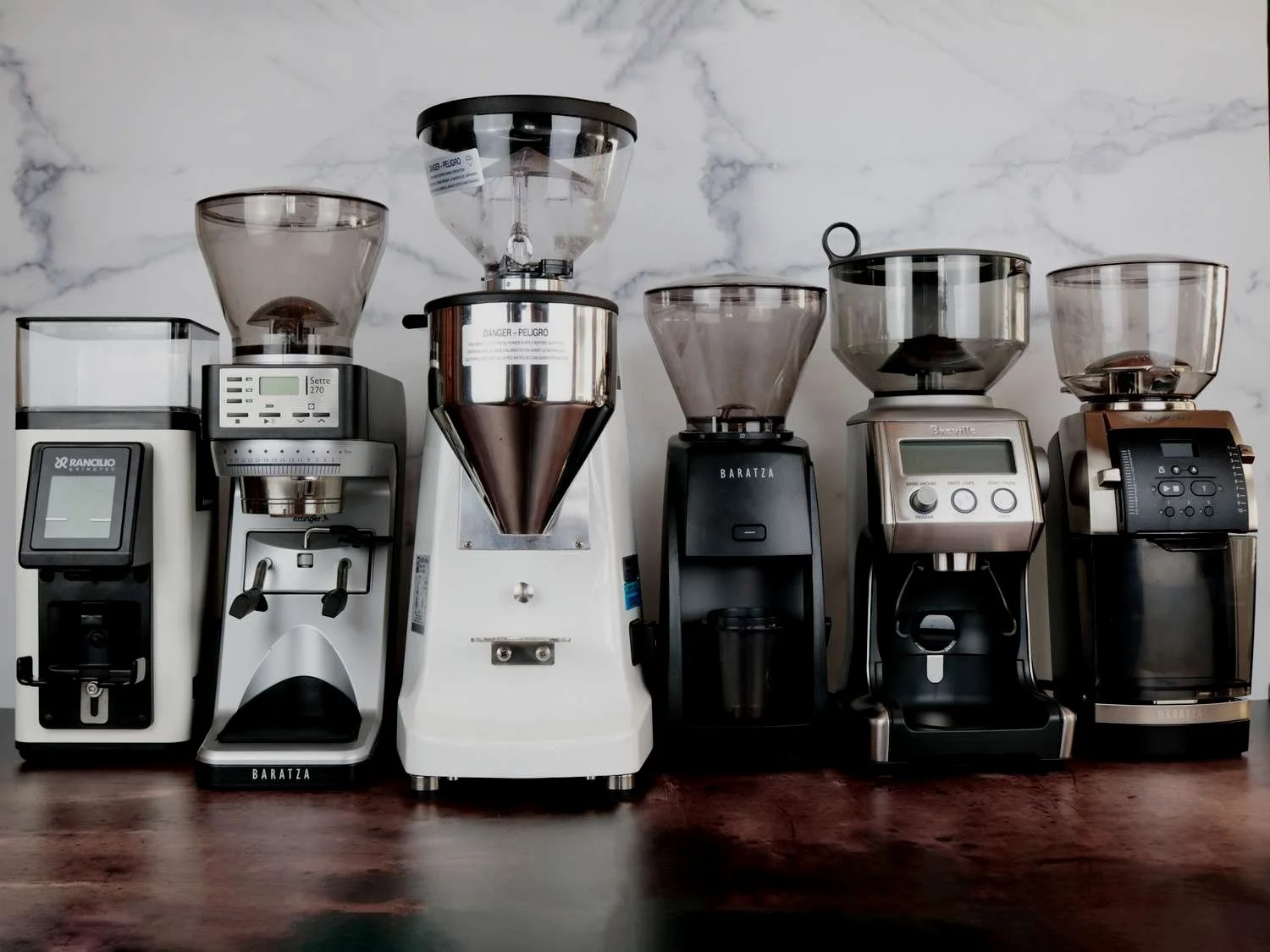 Line of six coffee grinders on a wooden surface with a marble wall background.