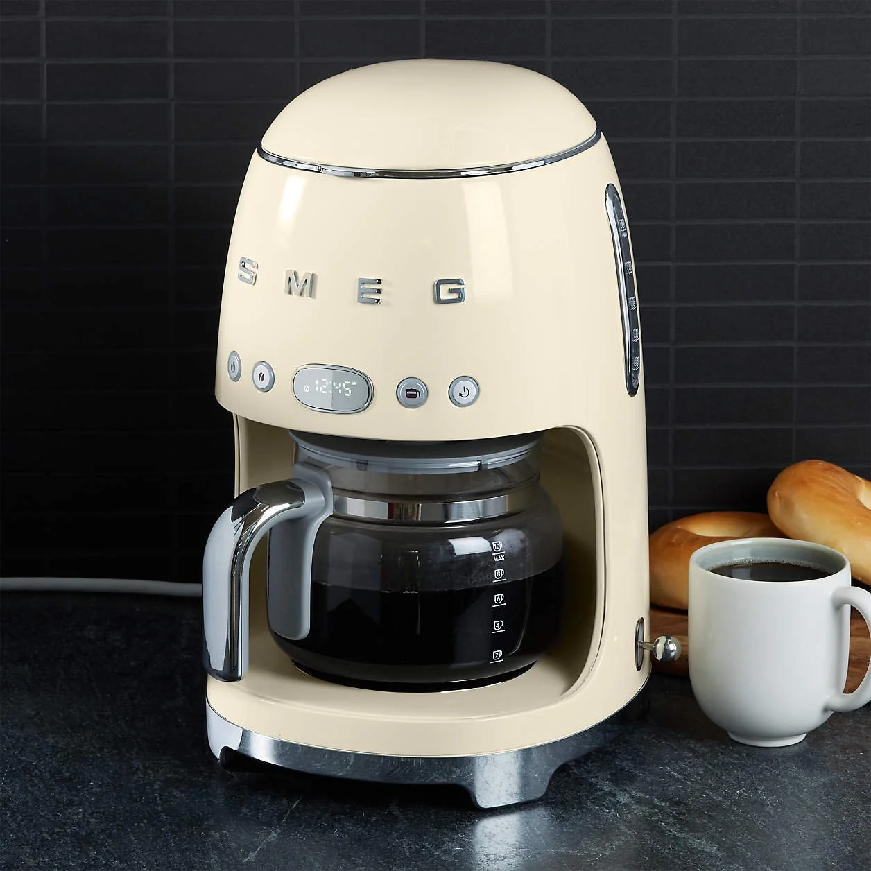 Cream-colored Smeg coffee maker next to a white coffee mug filled with black coffee and some bread rolls on a dark countertop.