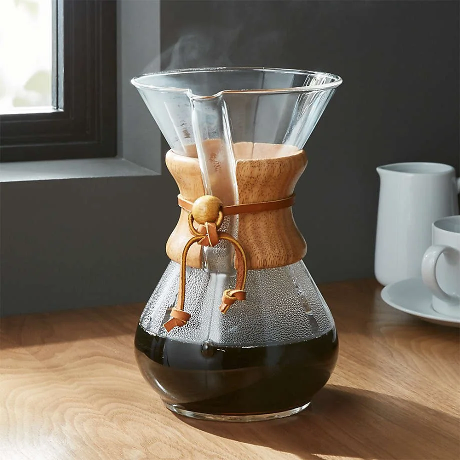 Glass Chemex coffee maker with brewed coffee on a wooden table near a window and white cups in the background.