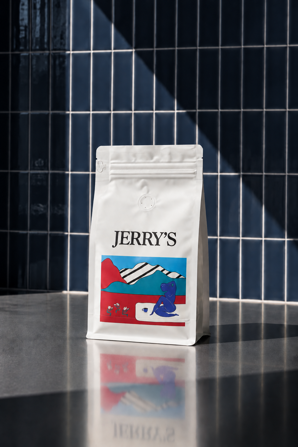 White coffee bag labeled 'Jerry's' with colorful mountains and a blue cartoon dinosaur illustration, placed on a reflective surface against a dark tiled wall background.