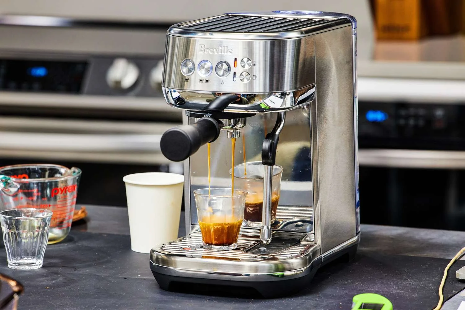 A stainless steel espresso machine brewing two shots of espresso into glasses, with coffee dripping into the cups. There are measuring cups and a white paper cup nearby on the black countertop.