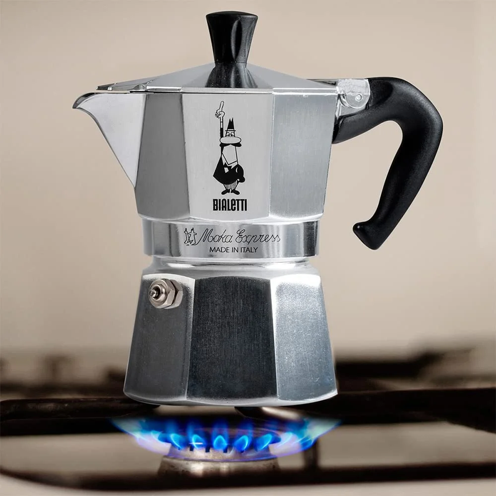 A moka pot coffee maker on a gas stove burner with blue flame, labeled Bialetti, Made in Italy.