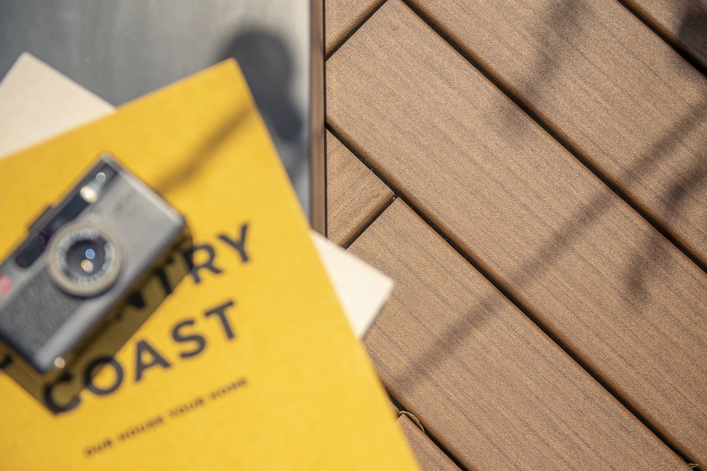 Close-up of a vintage camera on top of a yellow book titled 'COAST' with some papers underneath, next to a wooden slatted surface.