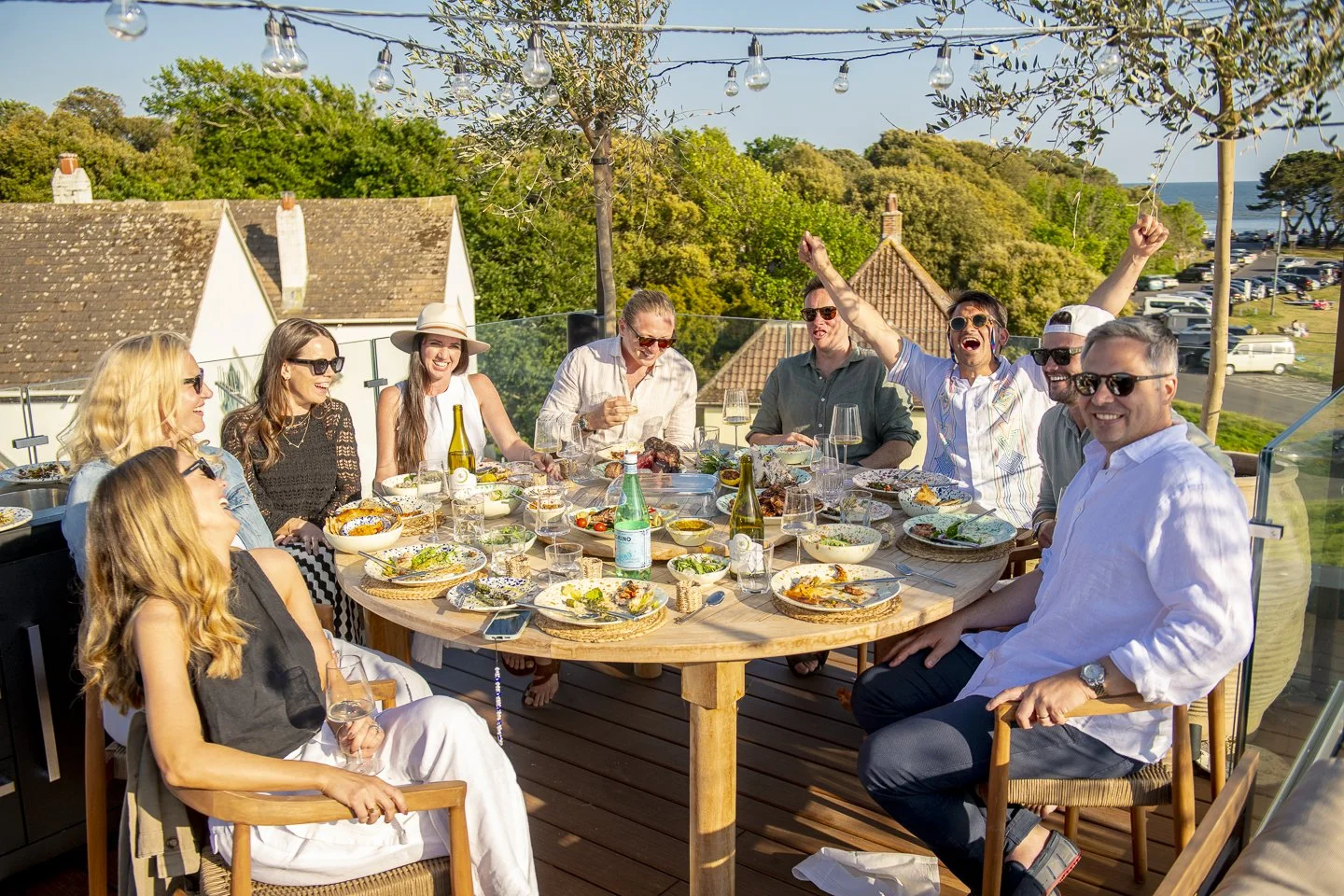 A group of people enjoying a meal outdoors on a decorated patio with string lights, surrounded by trees and buildings, overlooking the ocean.