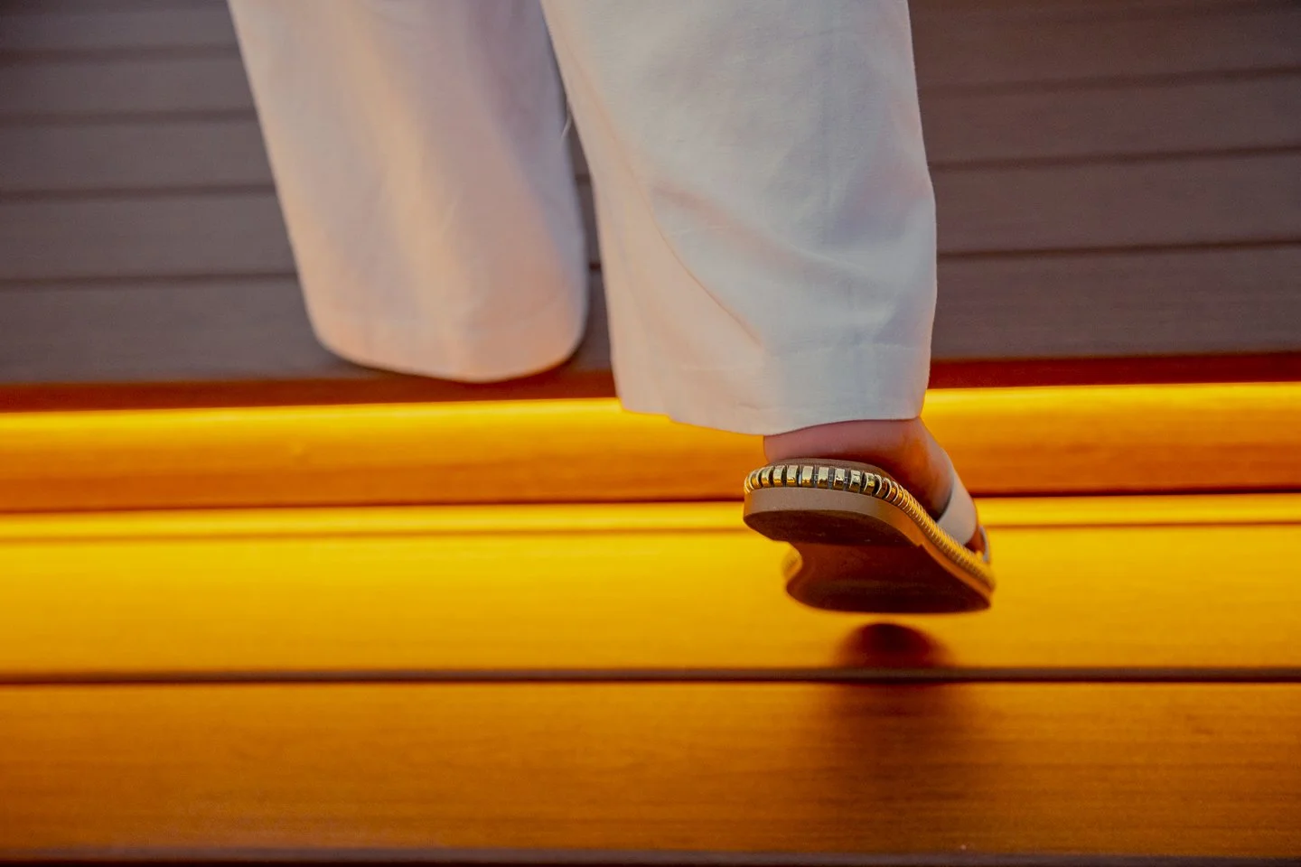 Close-up of a person's foot in a sandal with a beaded strap stepping onto a wooden surface illuminated with warm yellow light, against a background of horizontal wooden panels.