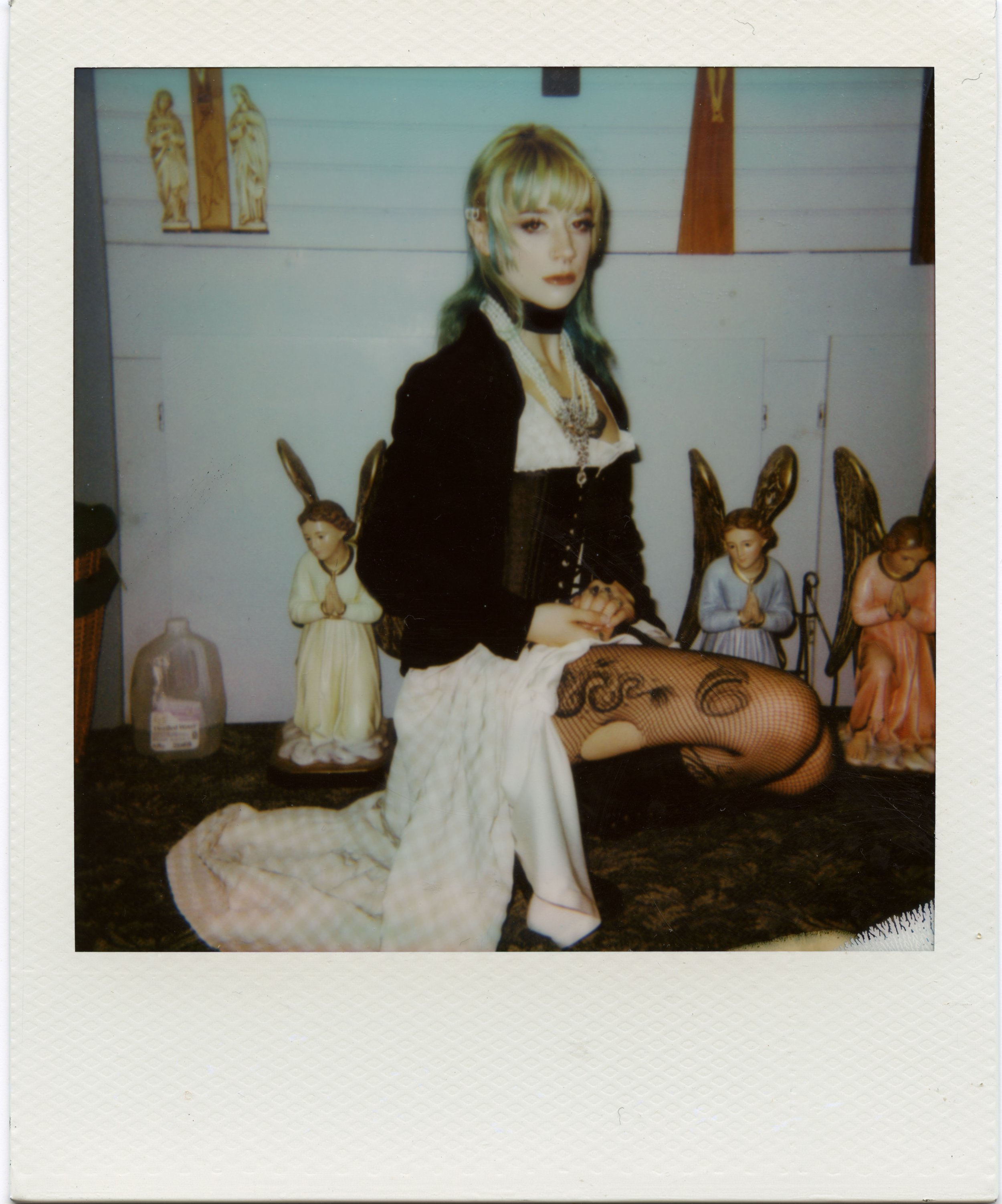 A woman with blonde hair and fishnet stockings seated in front of religious statues, with small figurines of angels and religious figures in the background.
