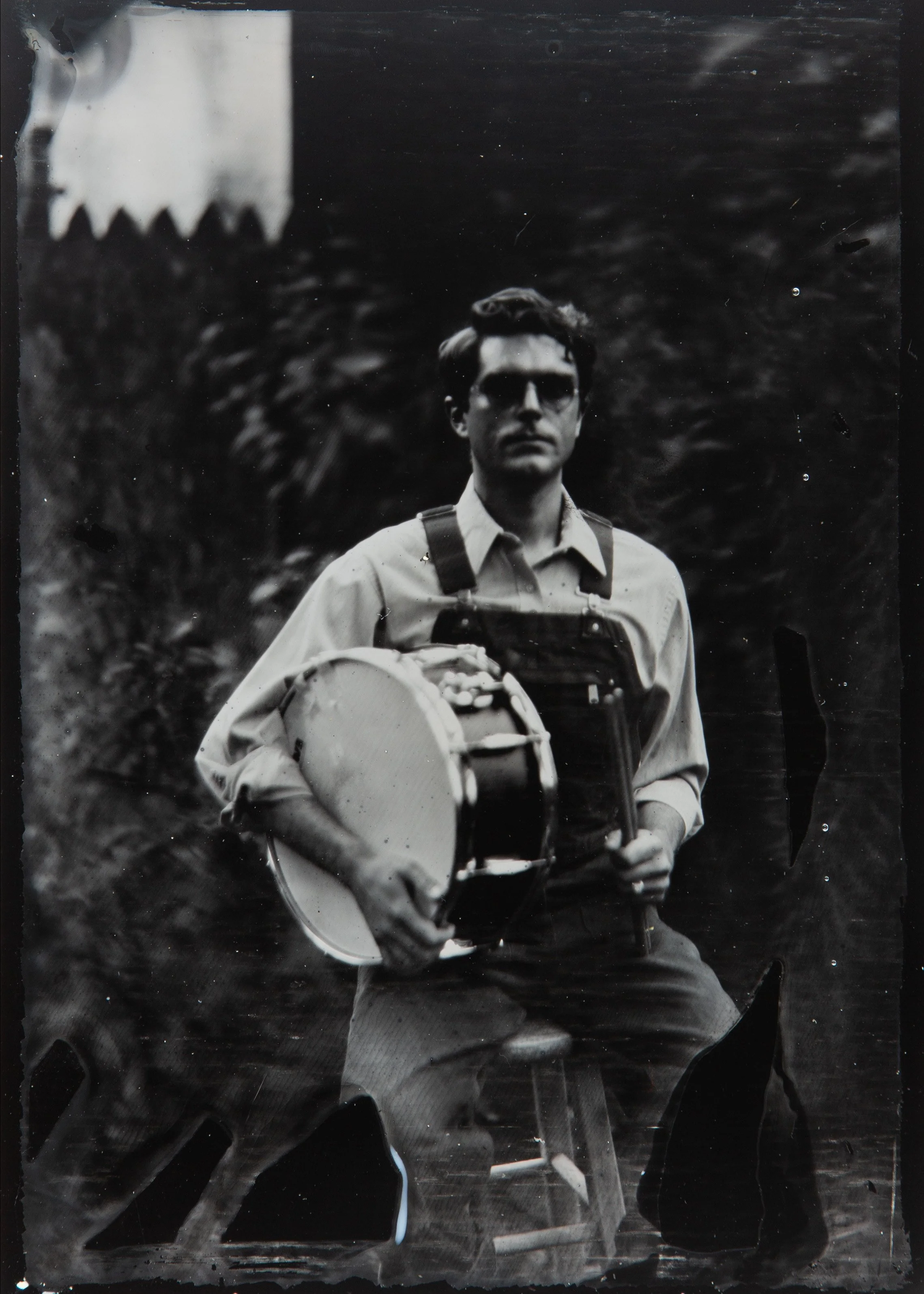 A black and white tintype photograph of a man with glasses and wavy hair, wearing a jacket, holding an instrument. The background is blurred with dark foliage, and there is a ladder or stairway in the lower part of the image.