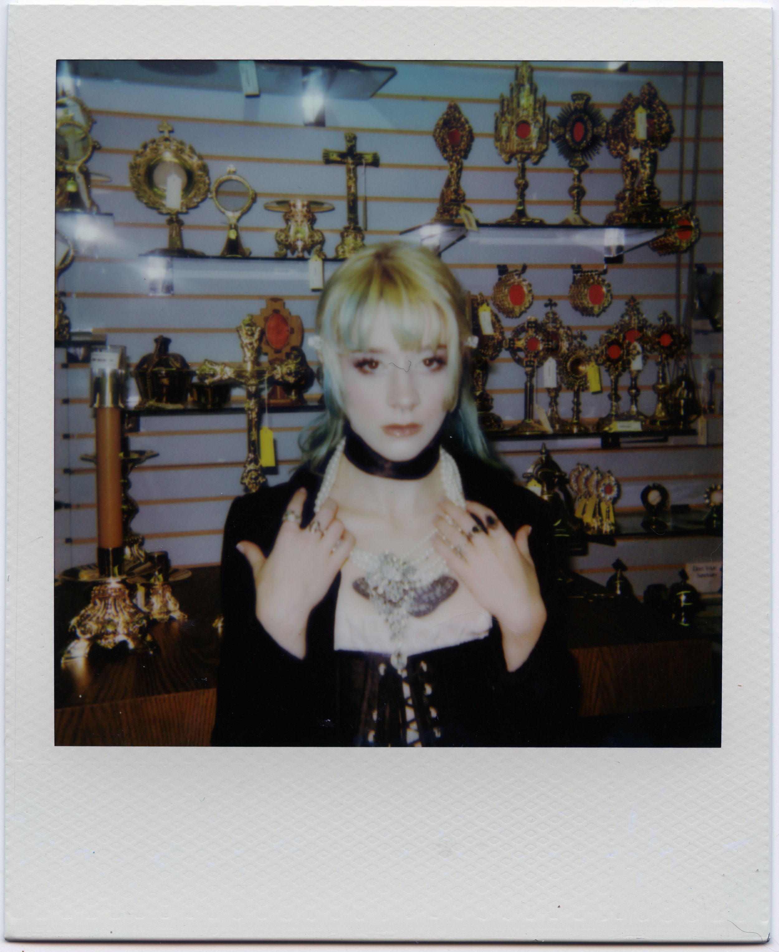 A person with green and blonde hair and dark makeup, wearing a black top and ornate jewelry, standing in front of shelves with gold and religious artifacts in a store.