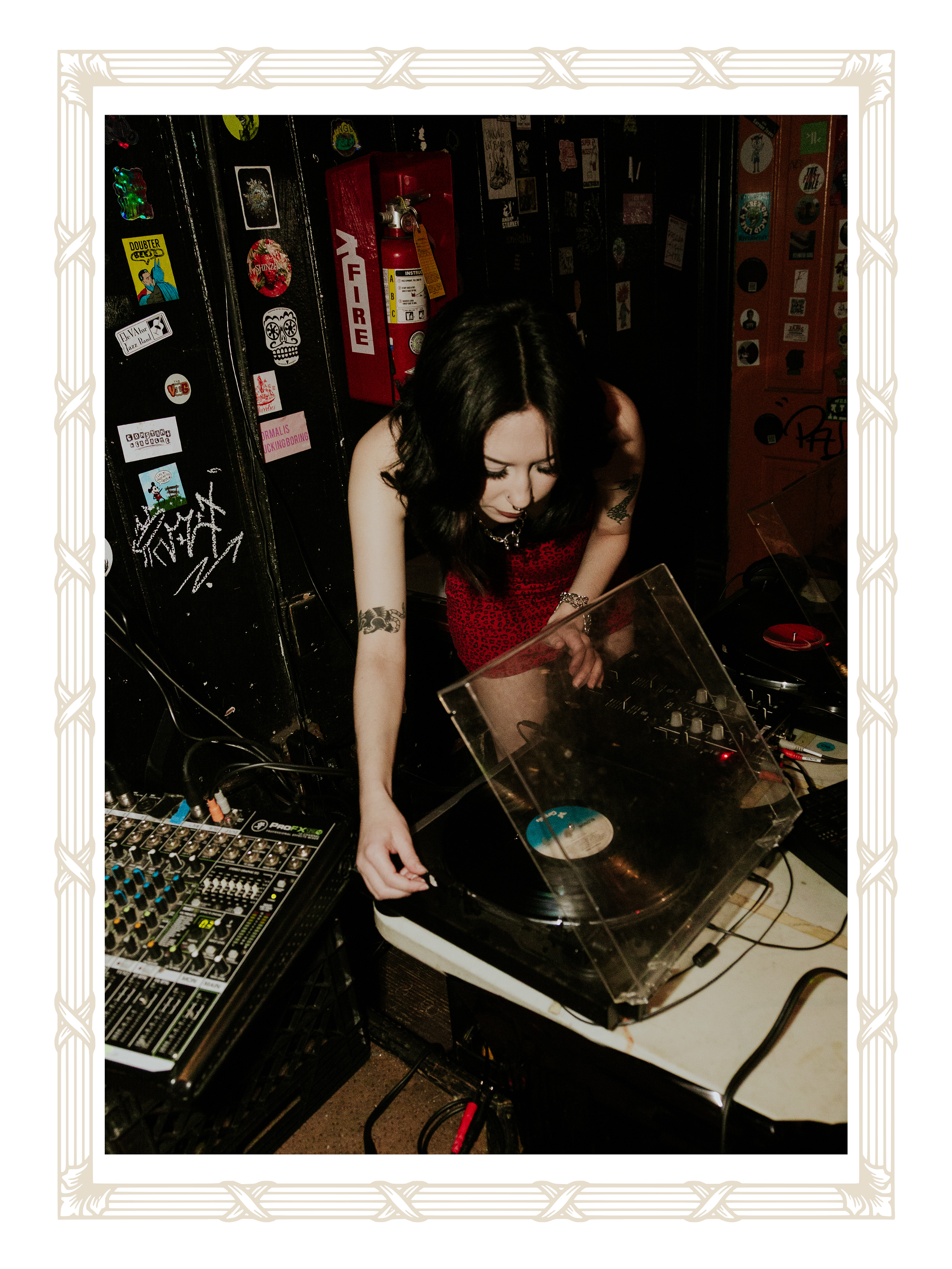 A woman with dark hair and tattoos, wearing a red top, is DJing with a turntable. She is leaning over the equipment in a room with black lockers covered in stickers, a red fire extinguisher, and electronic music gear.