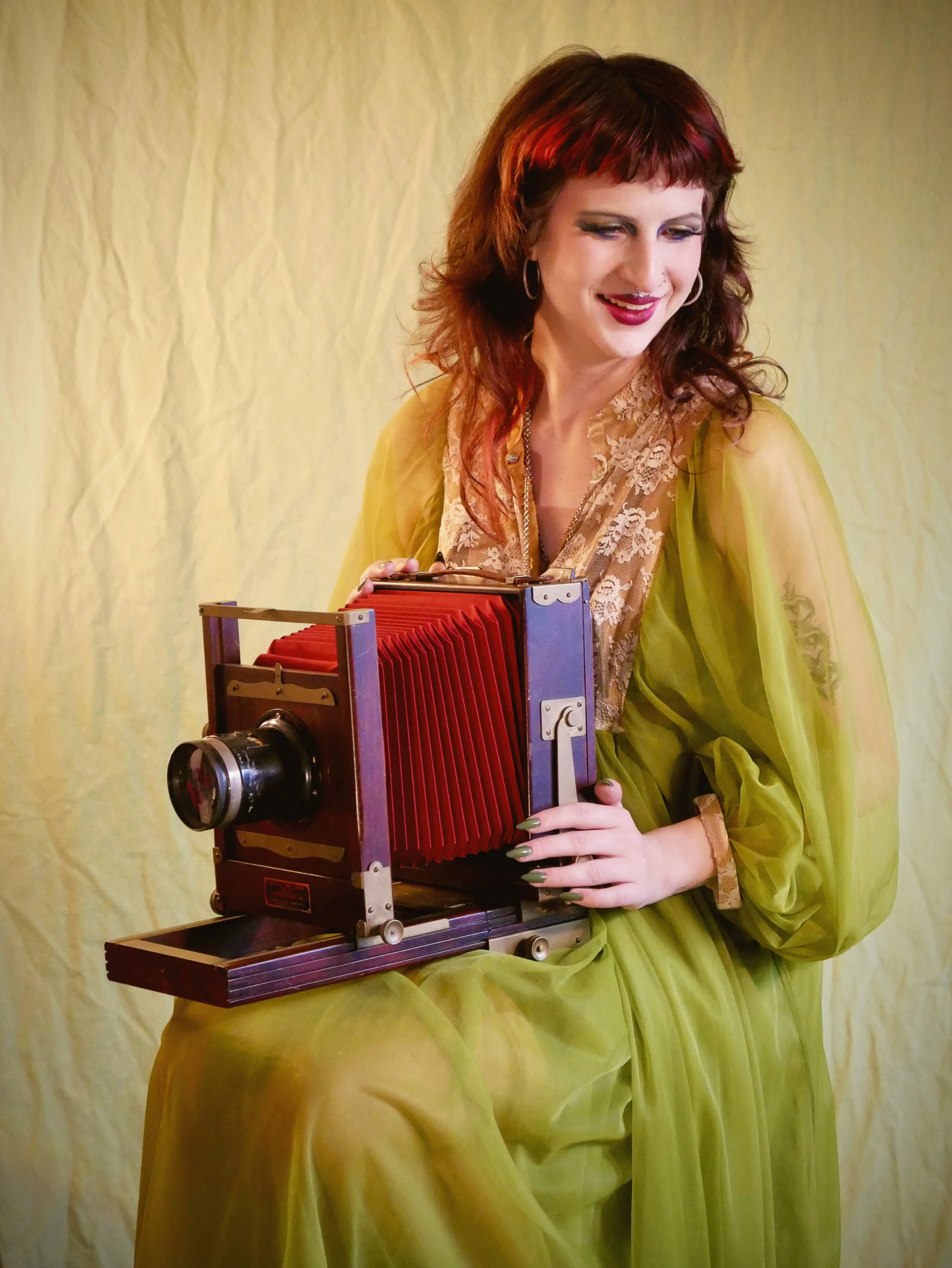 A woman with red hair, wearing a yellow dress and lace top, holding a vintage camera with red bellows, standing in front of a yellow backdrop.
