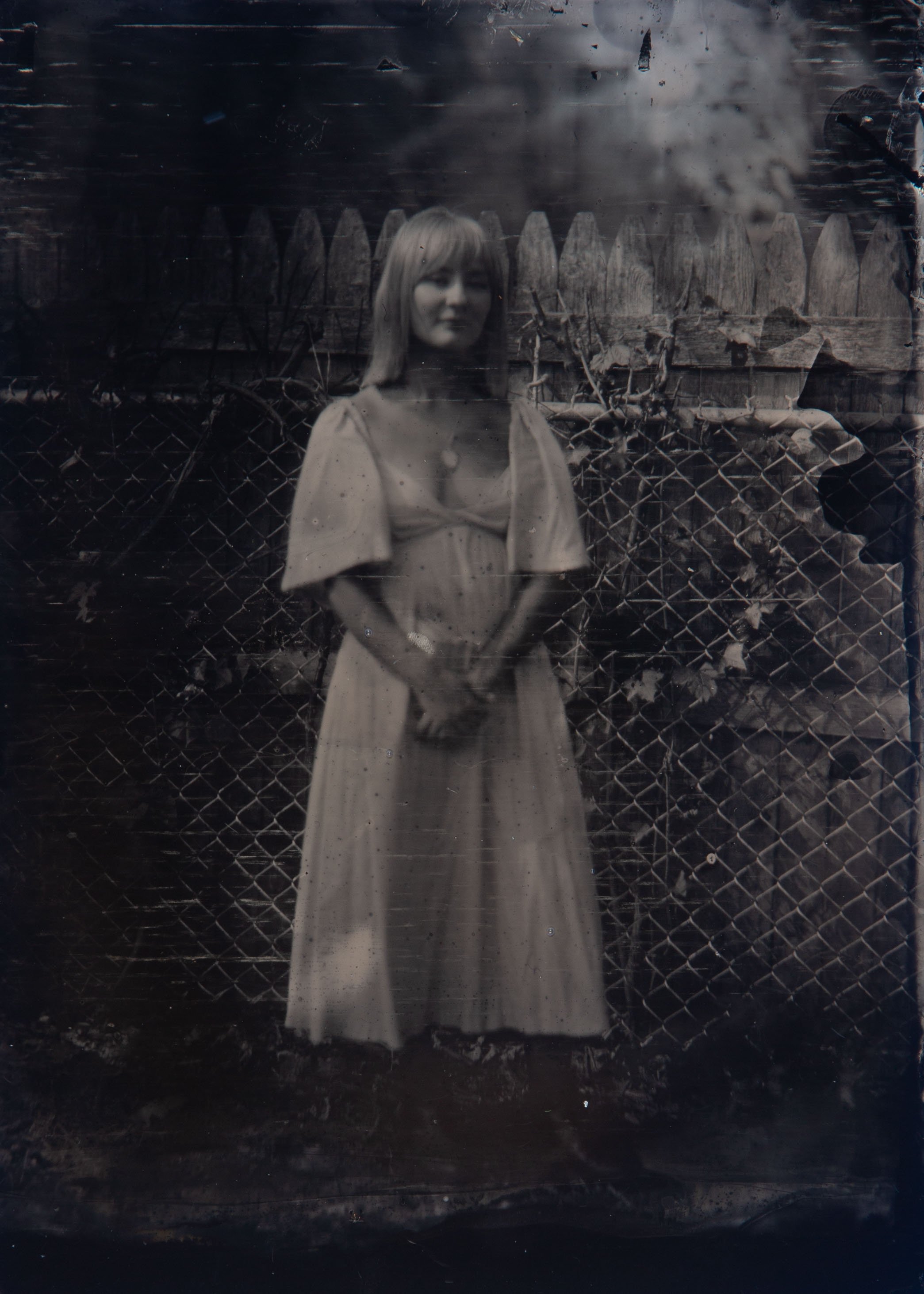 A black and white photo of a woman with long hair standing in front of a wooden fence with a chain-link fence at her feet. There are trees and clouds in the background.
