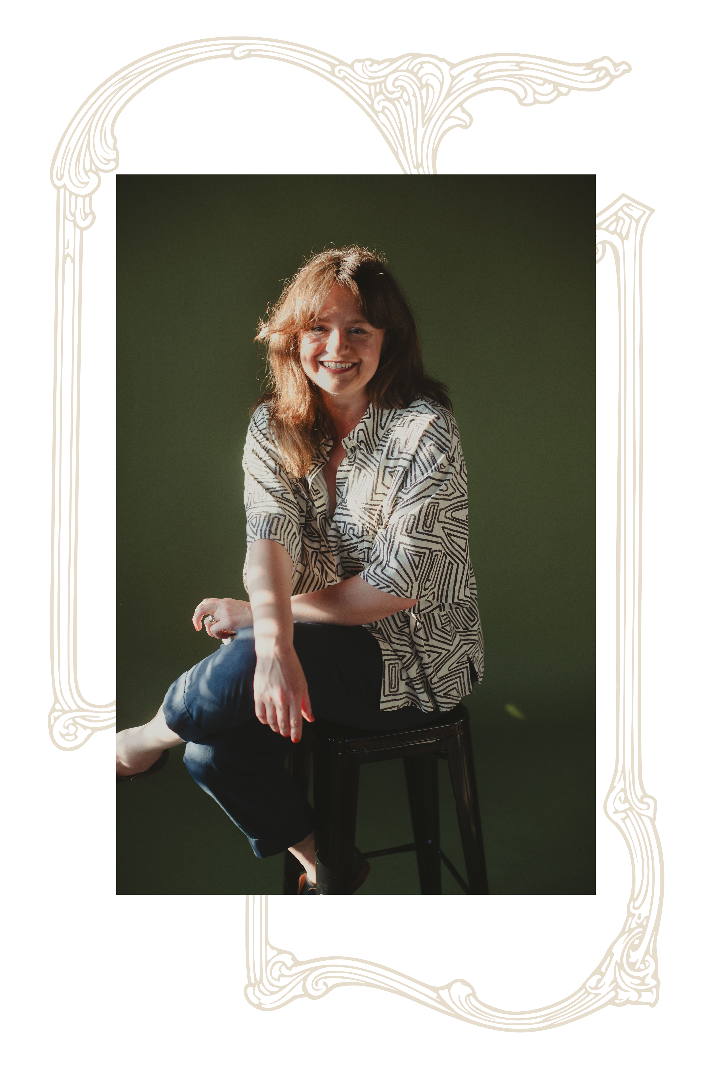 A young woman with shoulder-length light brown hair, smiling, sitting on a stool with one leg crossed over the other, wearing a patterned shirt and black pants, against a dark green background.