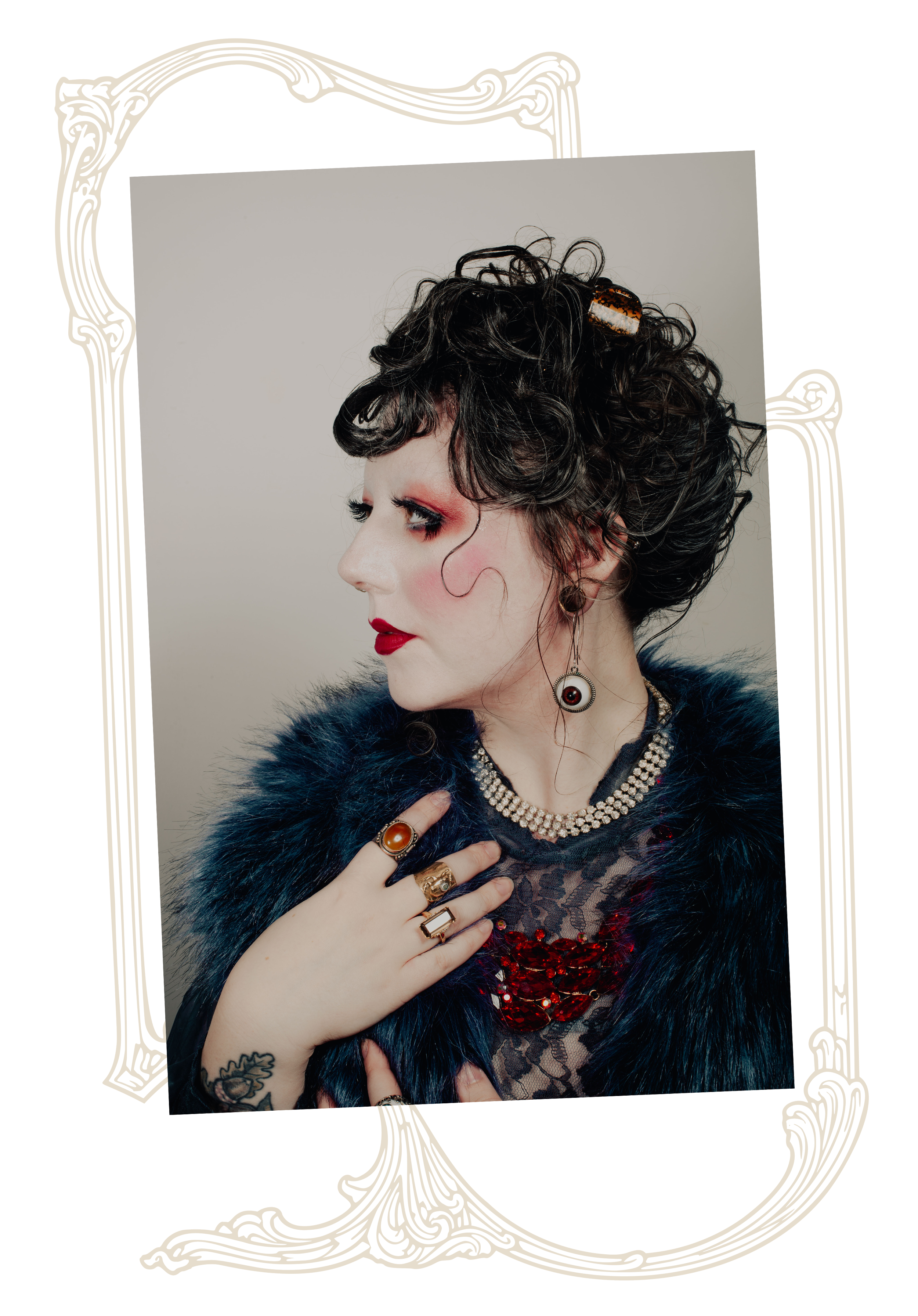 A woman with dark curly hair styled in a short, vintage look, wearing dramatic makeup with red lipstick and eye makeup, dressed in a lace top with red embellishments, accessorized with multiple rings, earrings, a pearl necklace, and a hair clip, with a dark fur coat over her shoulders, posing with her hand on her chest and looking away.