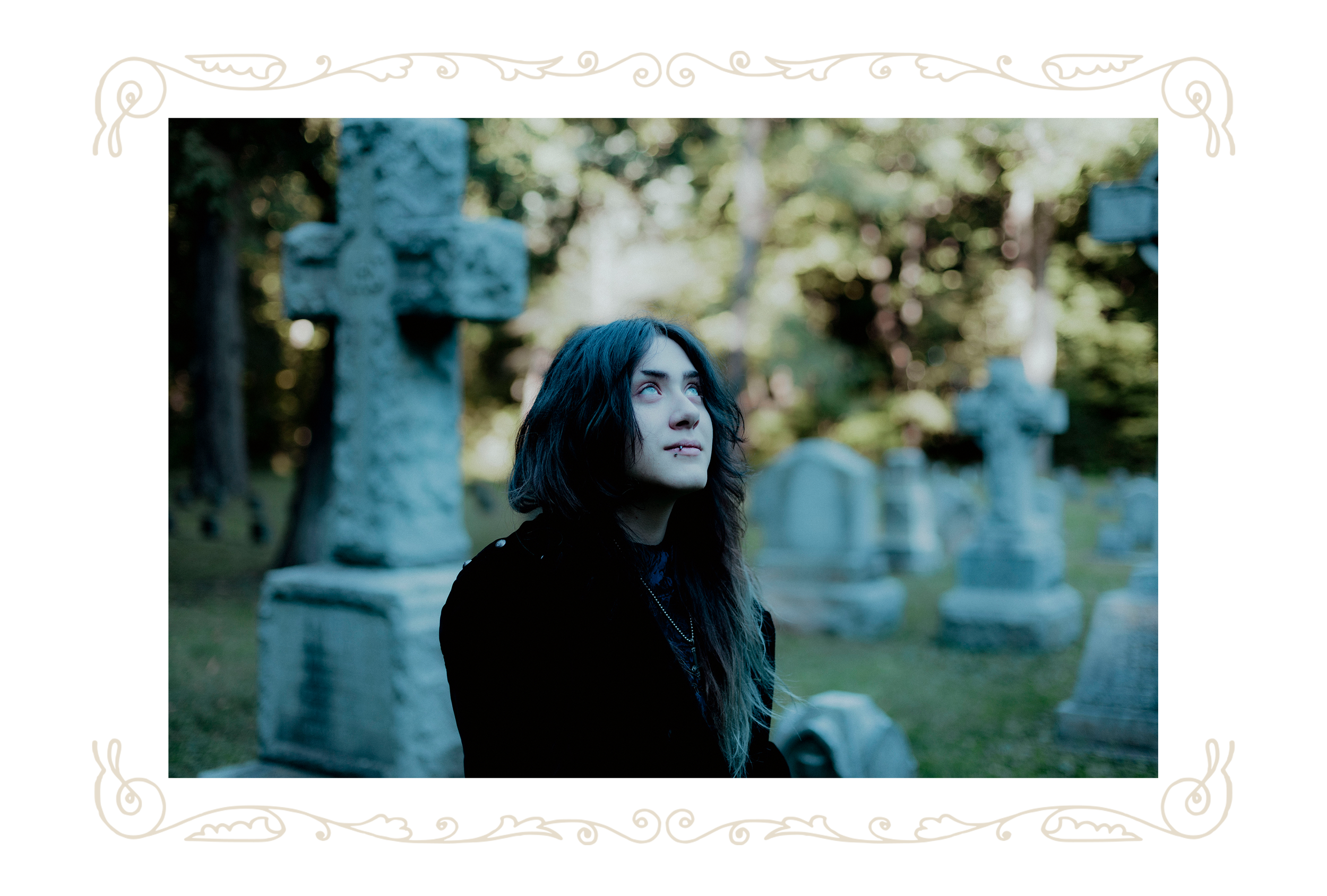 A young woman with long dark hair and piercings looking upwards in a cemetery with tombstones and a stone cross in the background.