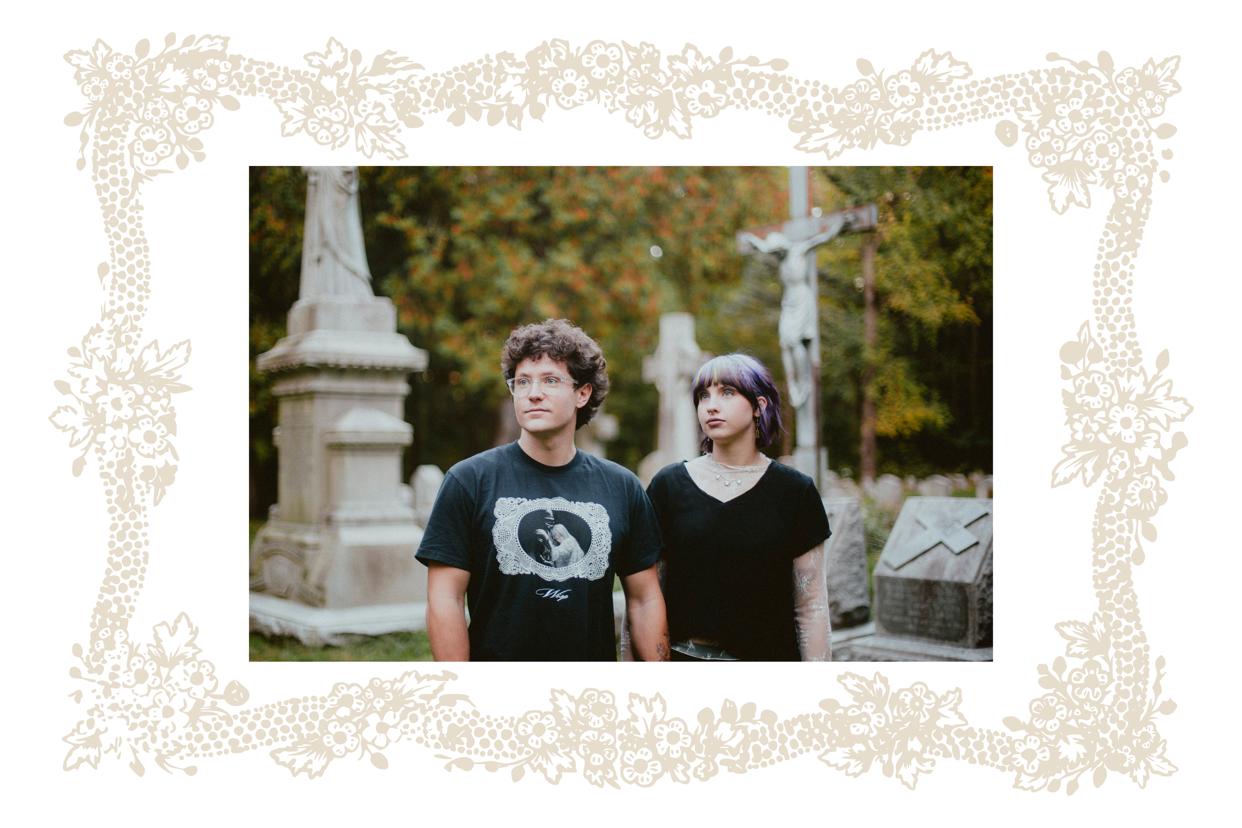Two young adults, a male with curly hair and glasses, and a female with shoulder-length hair with purple highlights, standing in a cemetery with tombstones and statues, surrounded by autumn trees.