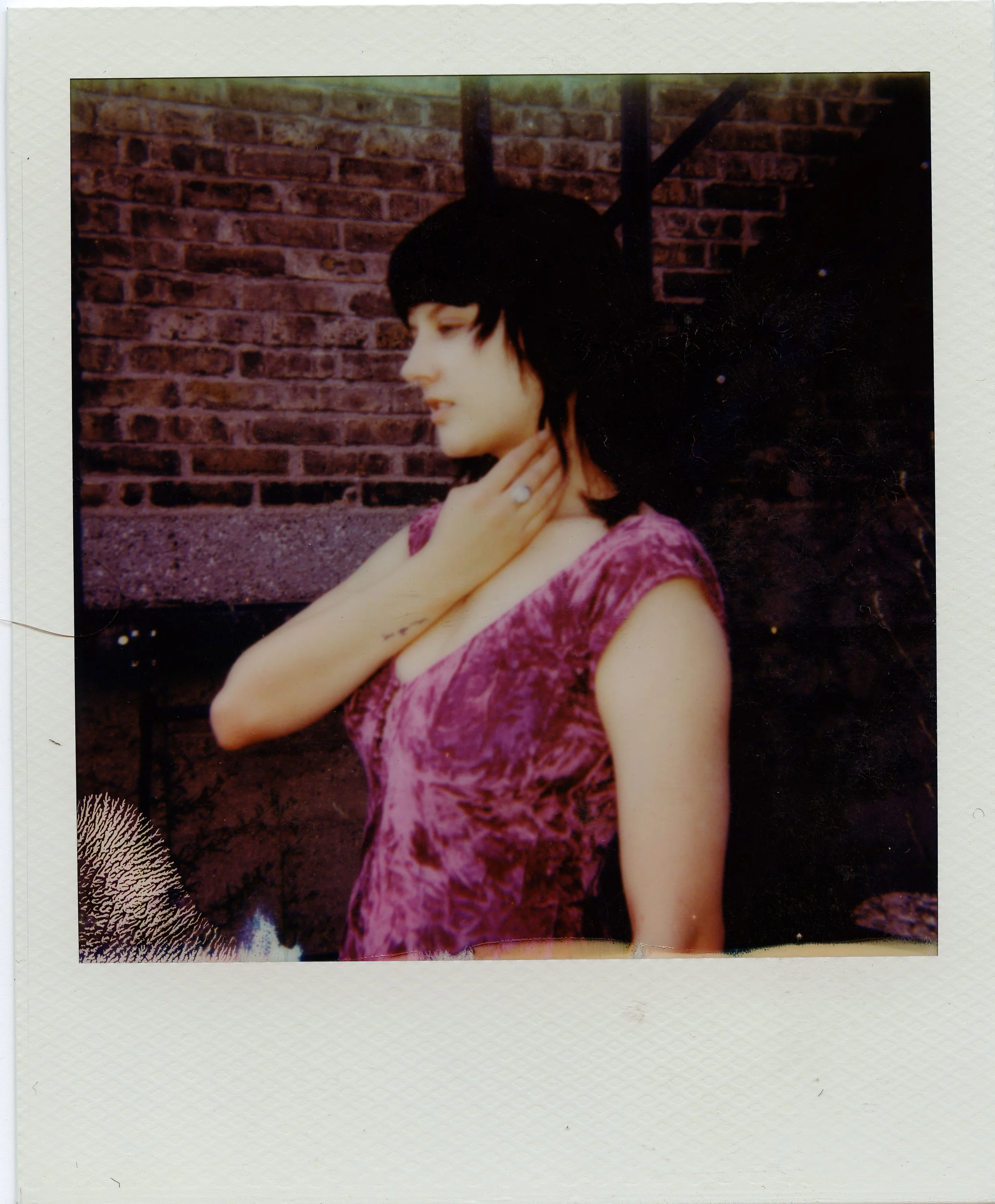A woman in a purple lace dress with black hair, standing outdoors against a brick wall, with her hand touching her neck and a ring on her finger.