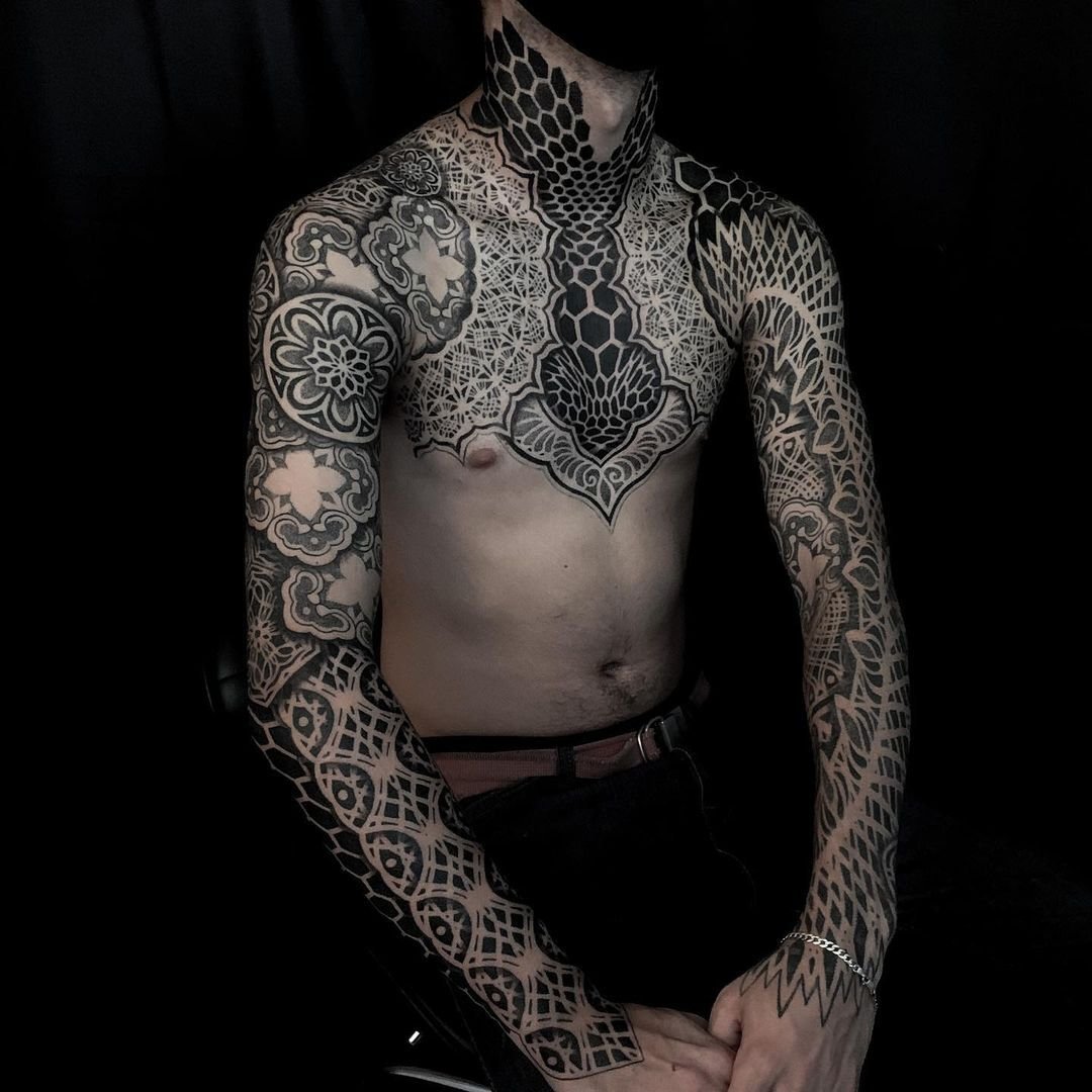 Man with intricate black tattoo designs on torso, arms, and neck against dark background.