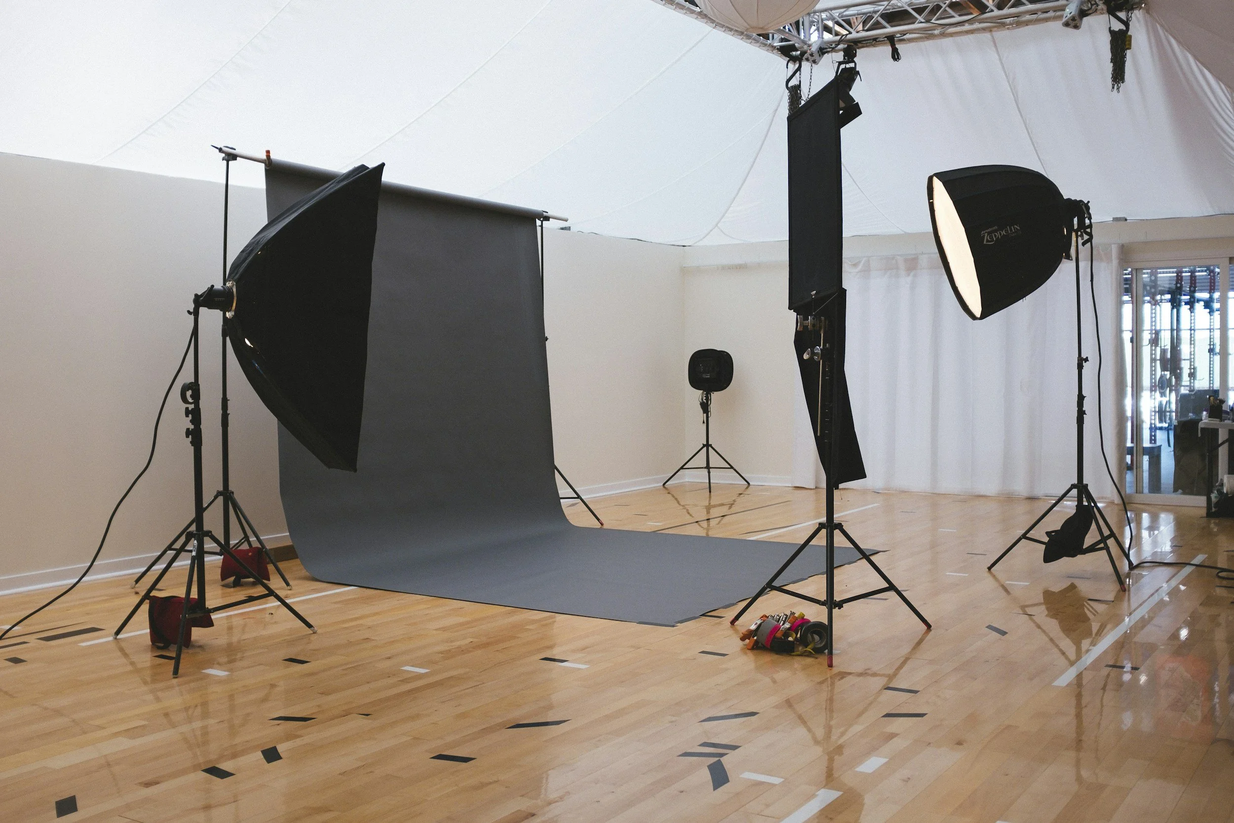 A professional photography studio setup with a gray seamless backdrop, mounted lights with softboxes, and a camera on a tripod. The studio has wooden floors, white curtains, and is indoors.