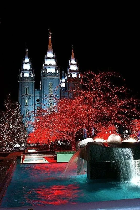 Nighttime view of a lit castle with three tall spires, surrounded by red-leaved trees and a fountain with cascading water in the foreground.