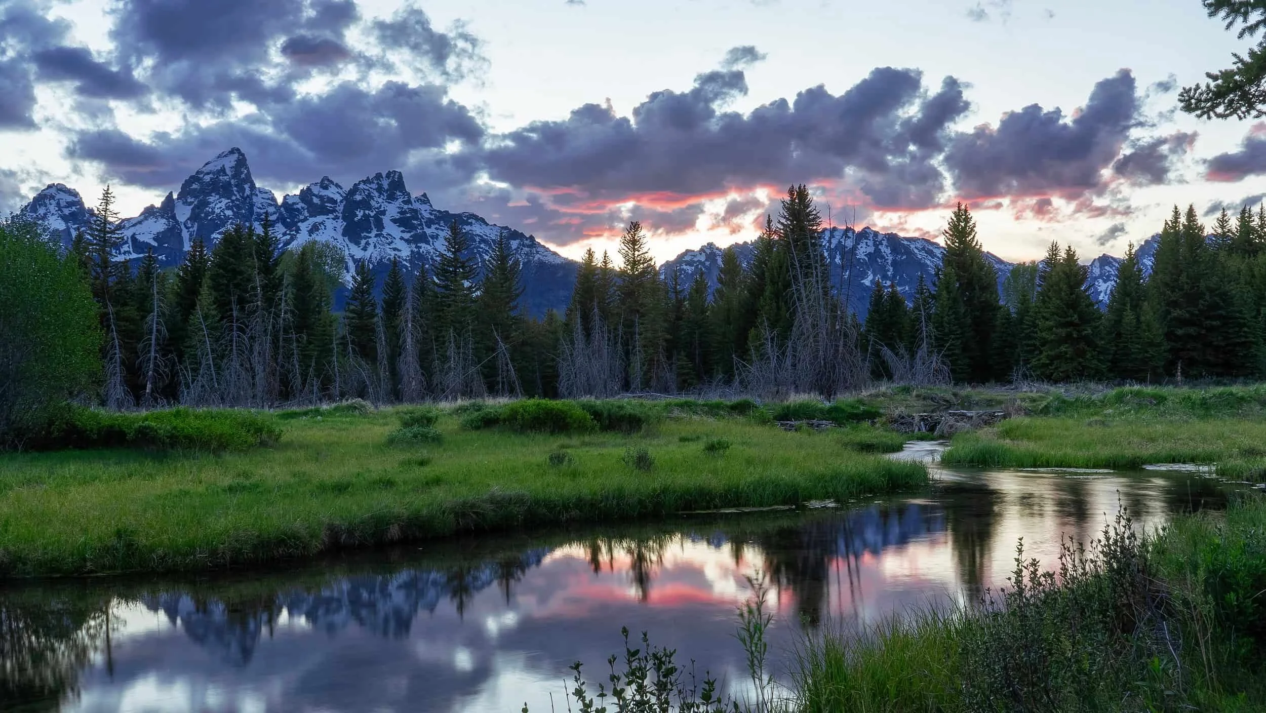 Snowy mountains and pine trees reflected in a calm stream at sunset—perfect for photography workshops amid a colorful sky and meadow.