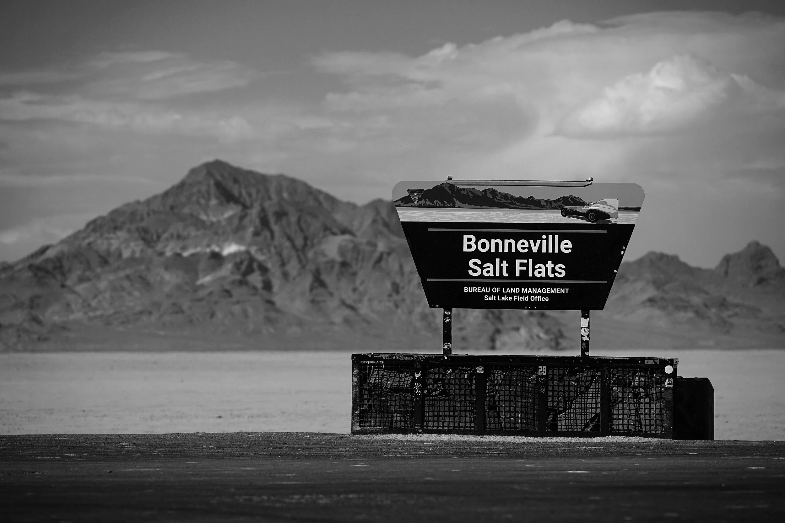 Black and white photo of a sign for Bonneville Salt Flats, with mountains in the background and a flat, expansive salt surface in the foreground