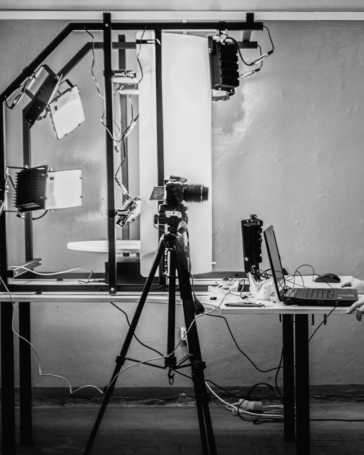 Photography setup with camera on tripod, laptop, and various electronic equipment on a table, behind a black metal frame with lights and wires in a studio or technical environment.