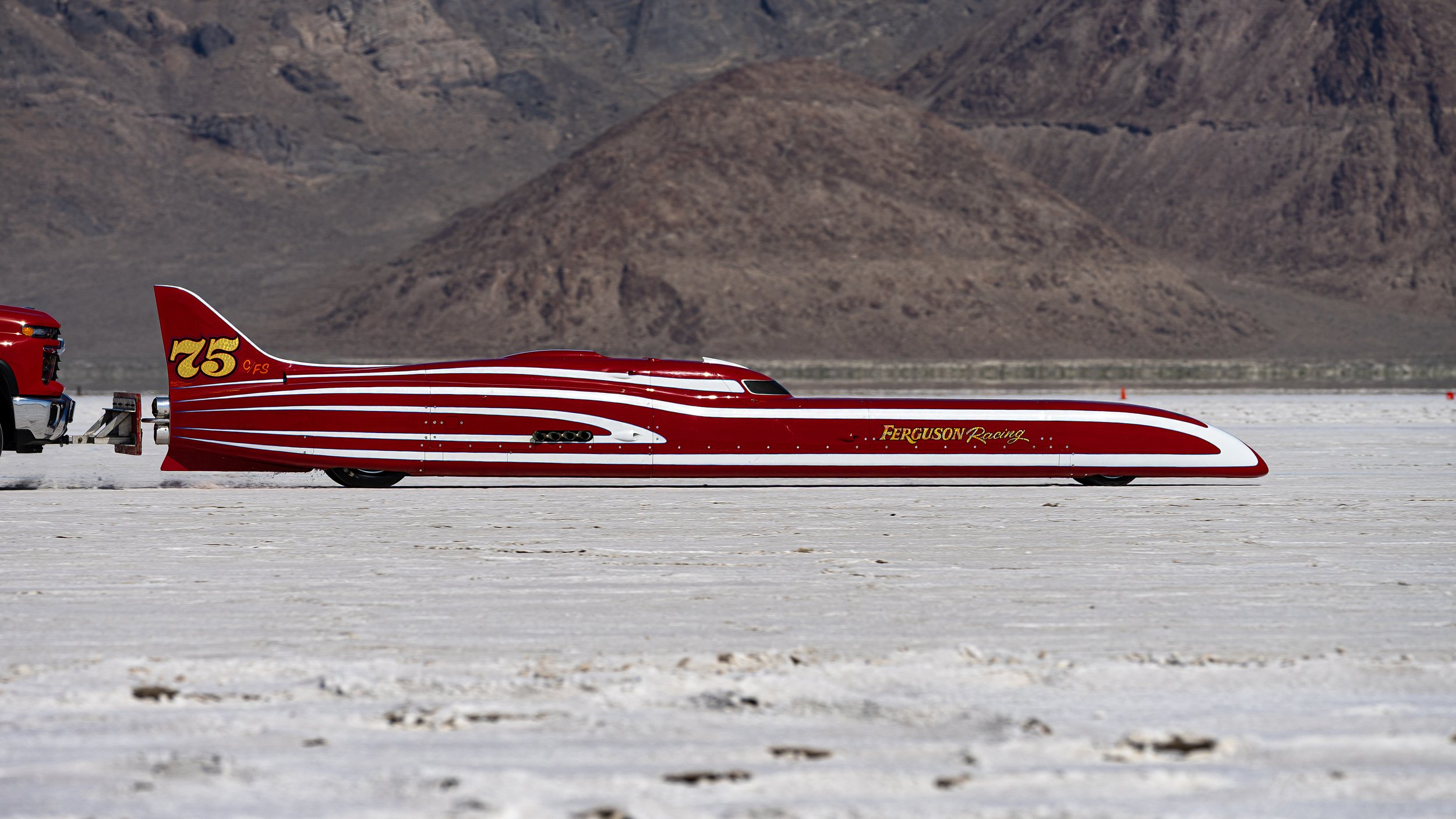 A red racing vehicle resembling a vintage jet or streamlined car with white stripes, displaying the number 75 and the words 'Ferguson Racing' on its side, on a flat white salt flat with mountains in the background.