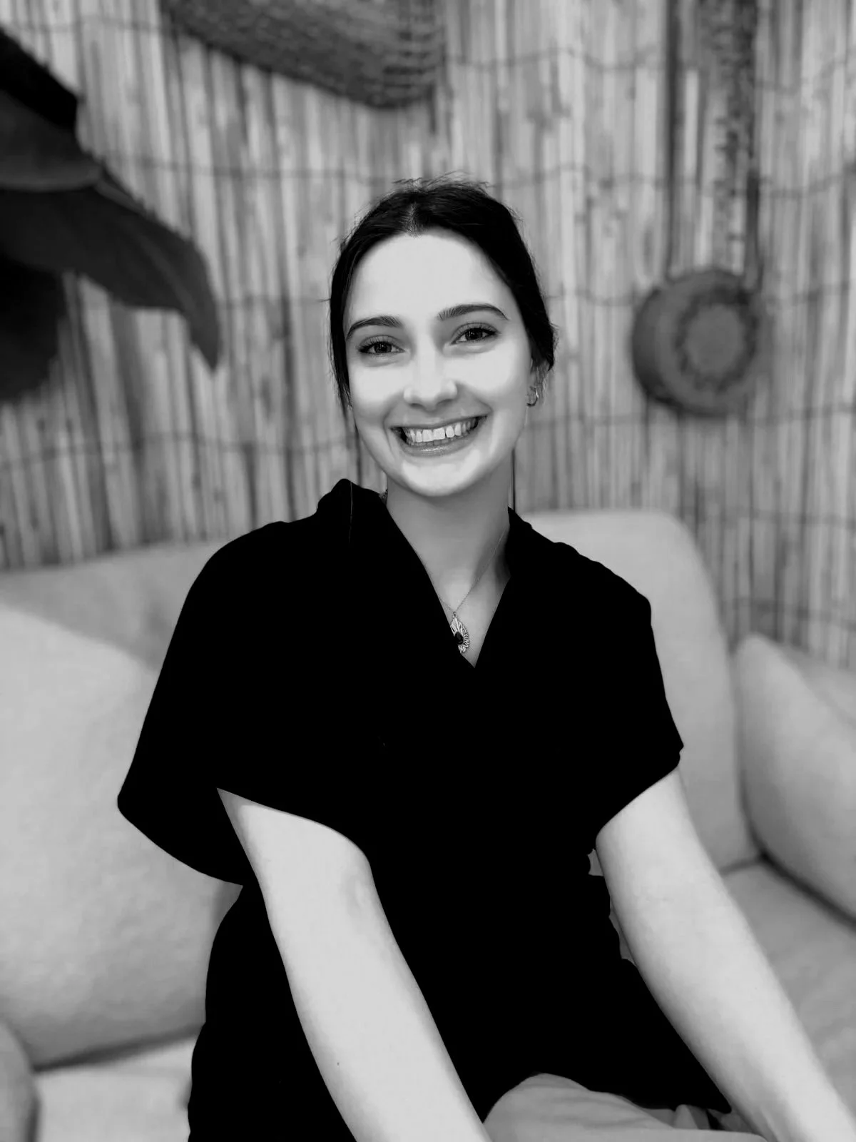 Un portrait en noir et blanc d'une femme souriante avec des cheveux bruns, portant un haut noir, assise dans un décor intérieur avec des éléments en bois et un tapis suspendu au plafond.